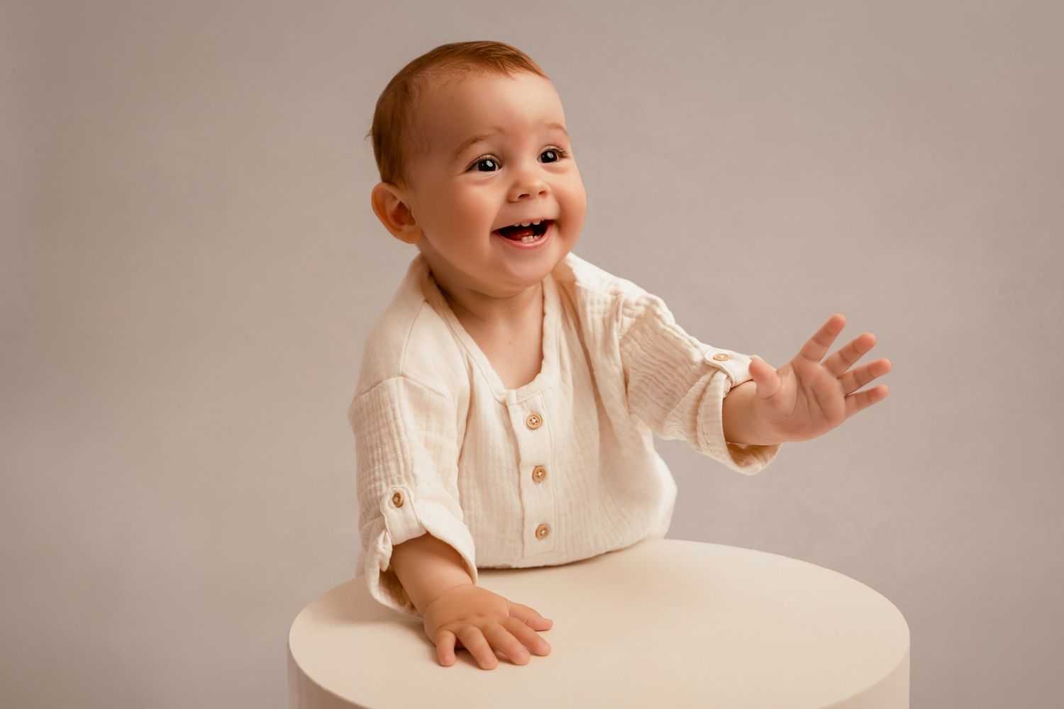Smiling baby in white linen outfit sitting at a white table, reaching out with one hand against a gray background.