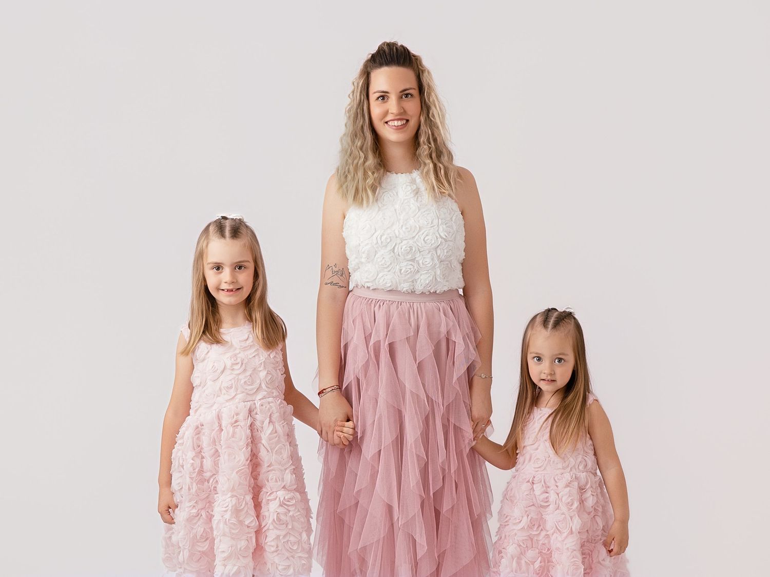 A mother in a pink tulle skirt holds hands with two young daughters in matching pink floral dresses on white background.