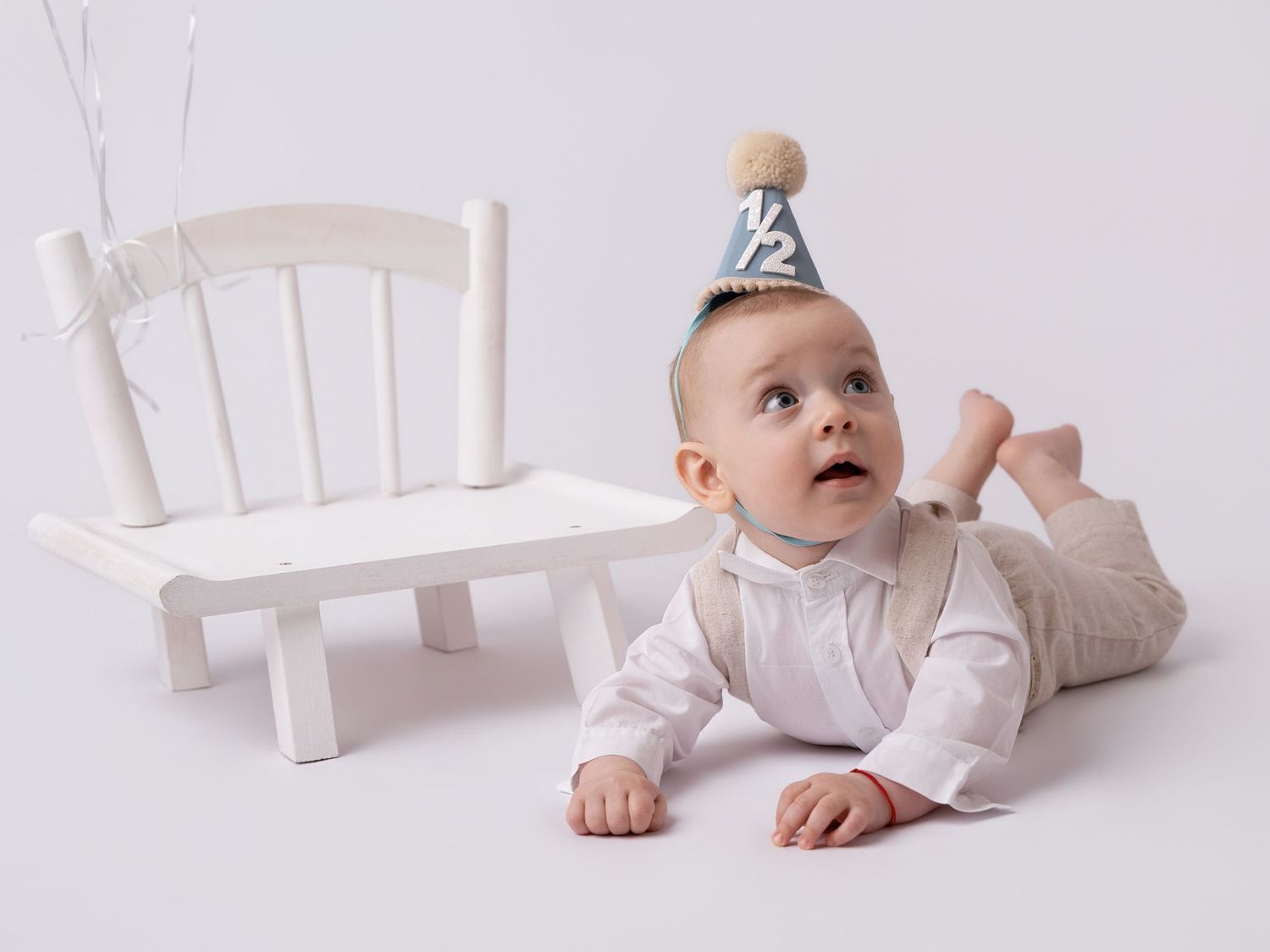 A baby wearing a half birthday party hat lies on their tummy on a white floor beside a small white chair.