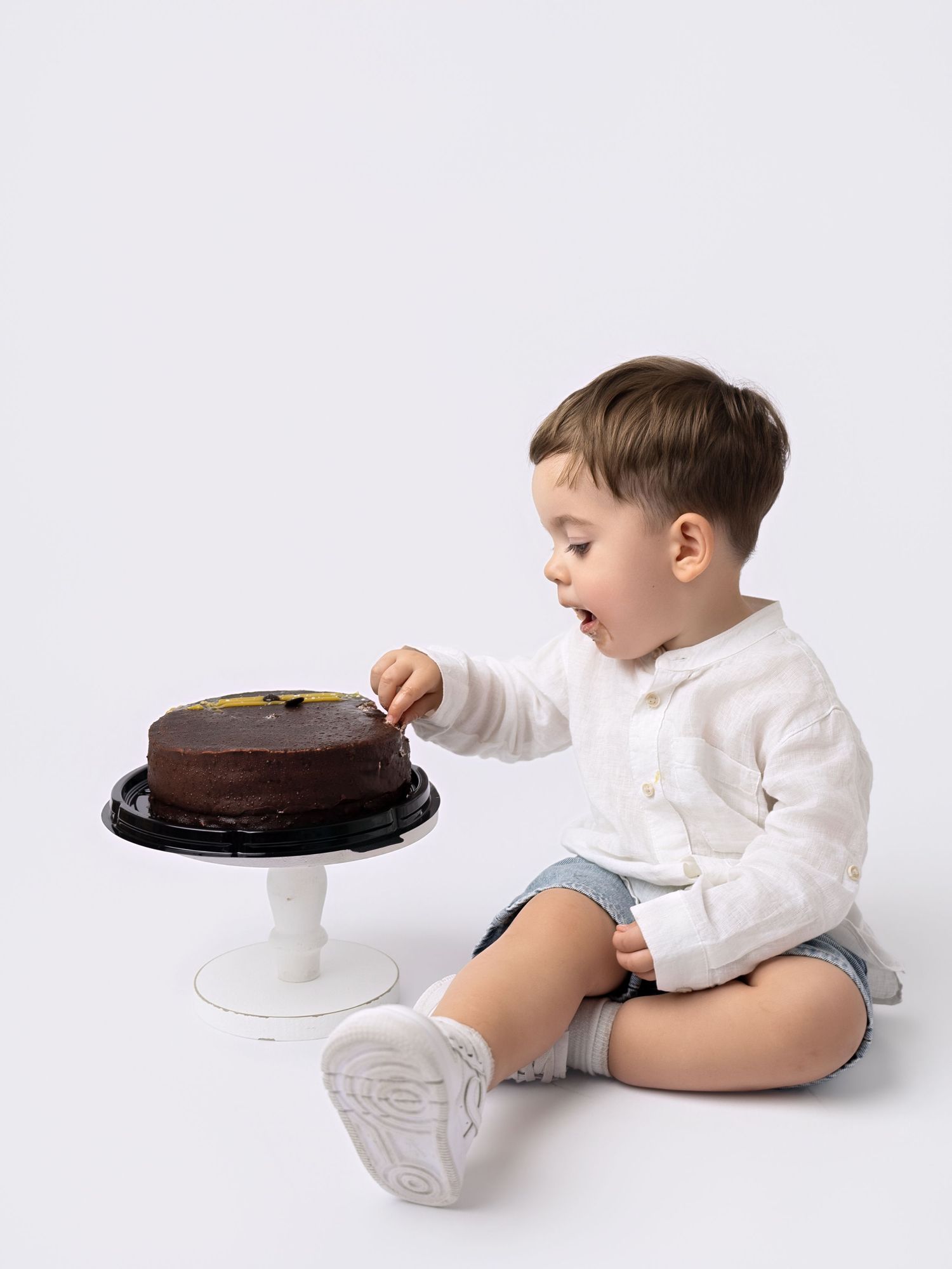 Baby boy in white shirt reaching to touch a chocolate cake on a white cake stand on white background.