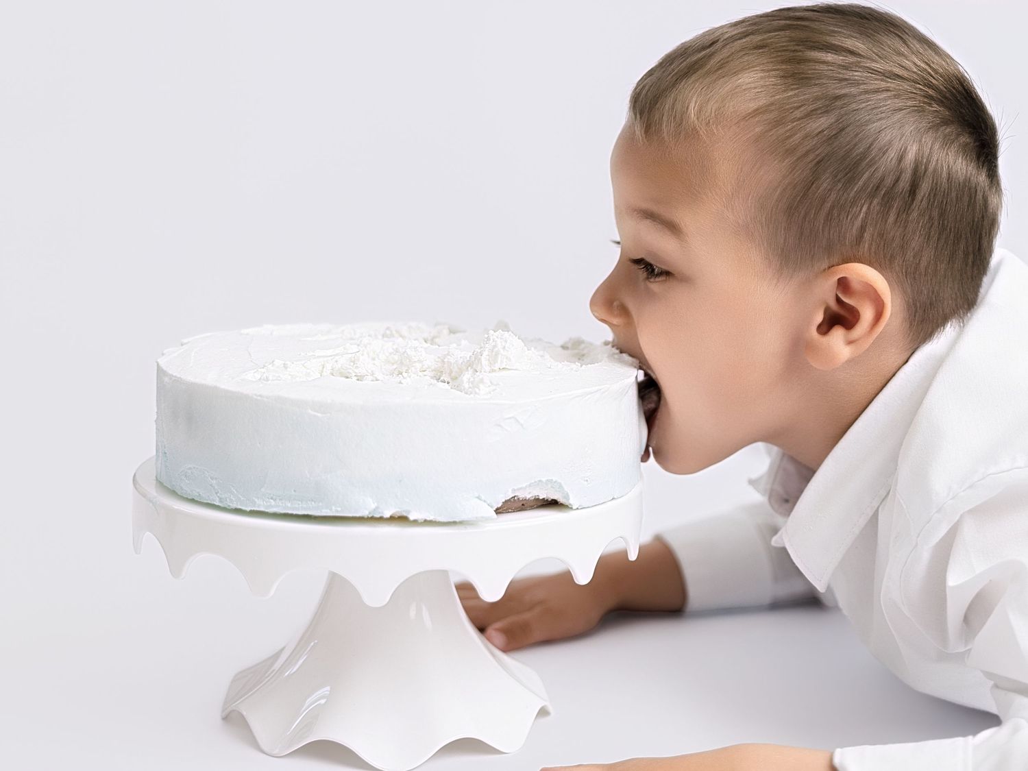 Young boy in white shirt biting into a white frosted cake on a cake stand against a white background.
