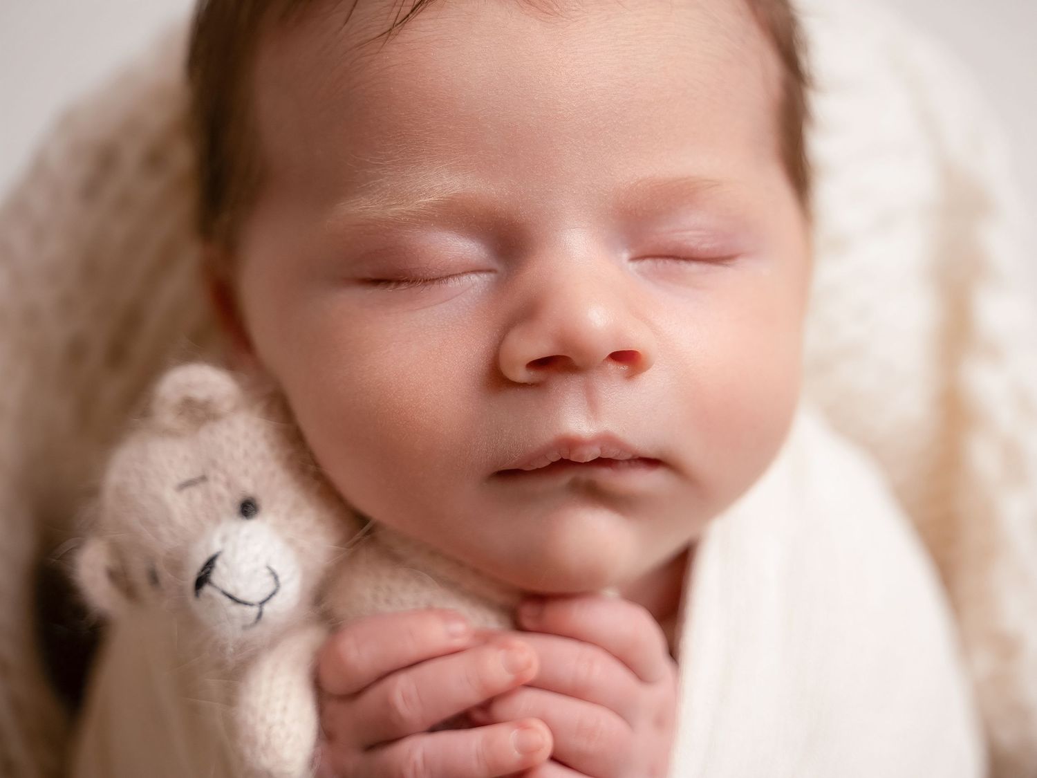Peaceful newborn baby sleeping with tiny hands clasped, cuddling a small knit teddy bear, wrapped in a soft cream blanket.