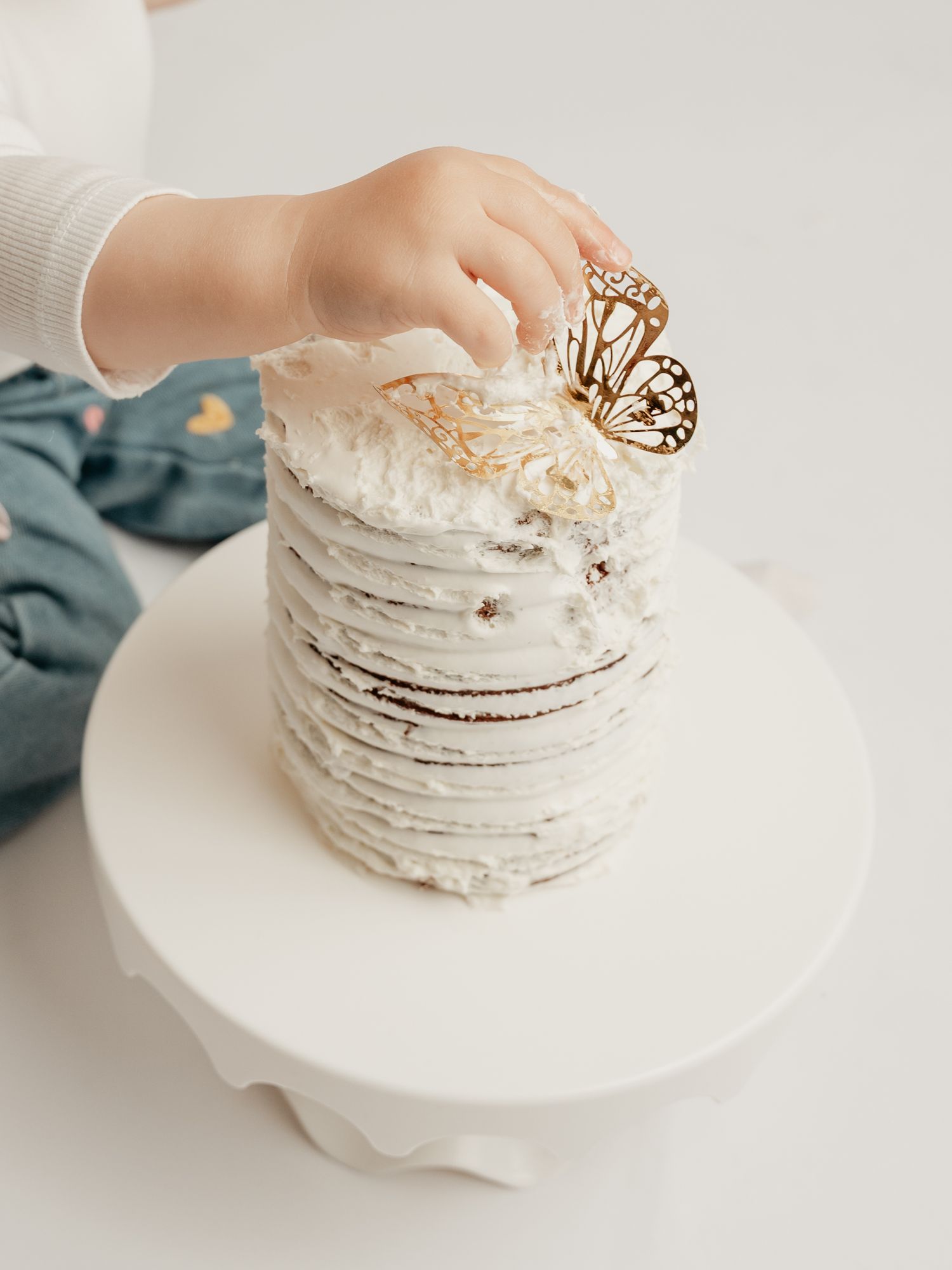 A toddler's tiny hand reaches for a gold butterfly decoration on a white layered cake.