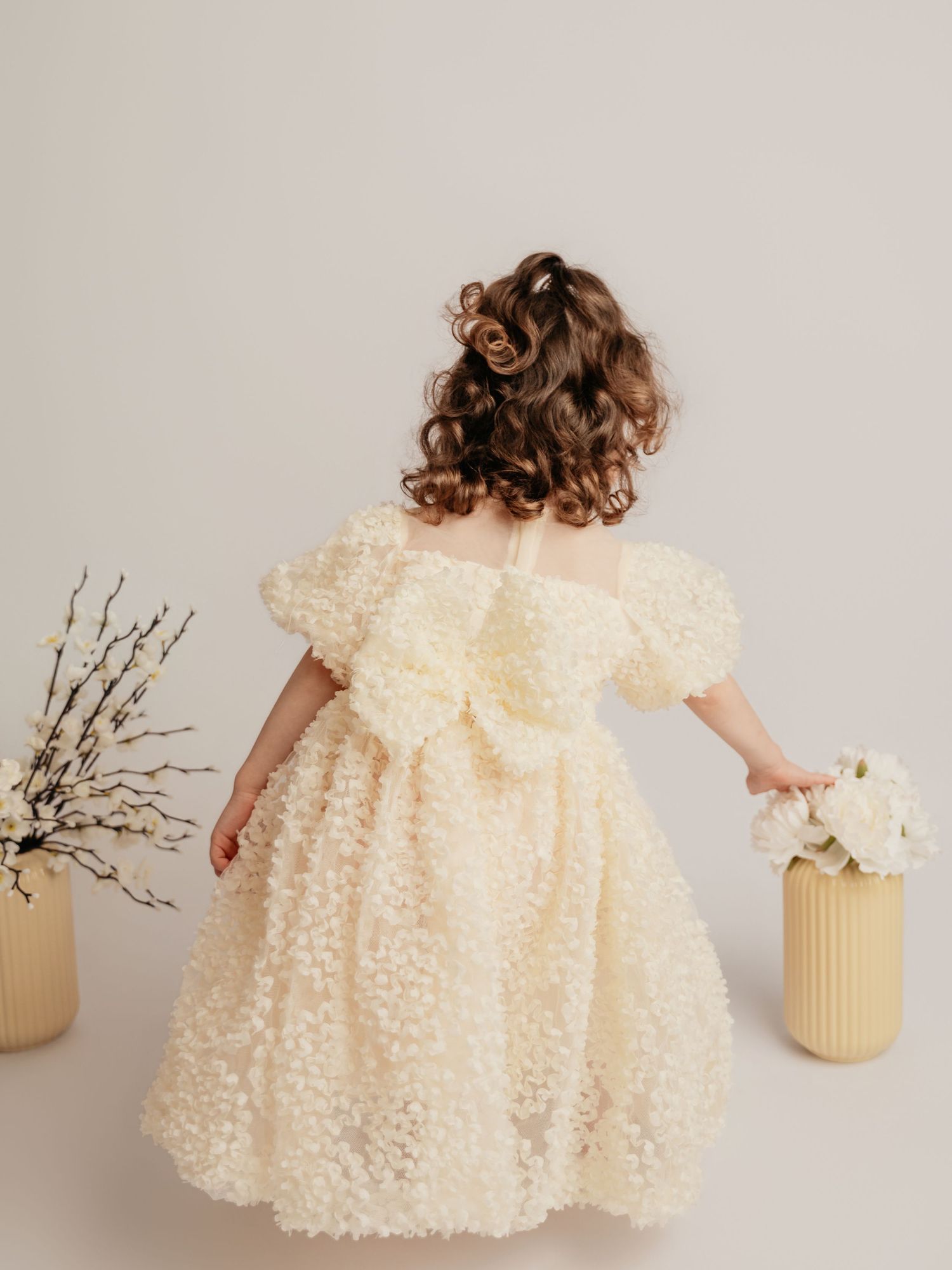 Back view of a young girl in a cream textured puff-sleeve dress with a large bow near floral vases.