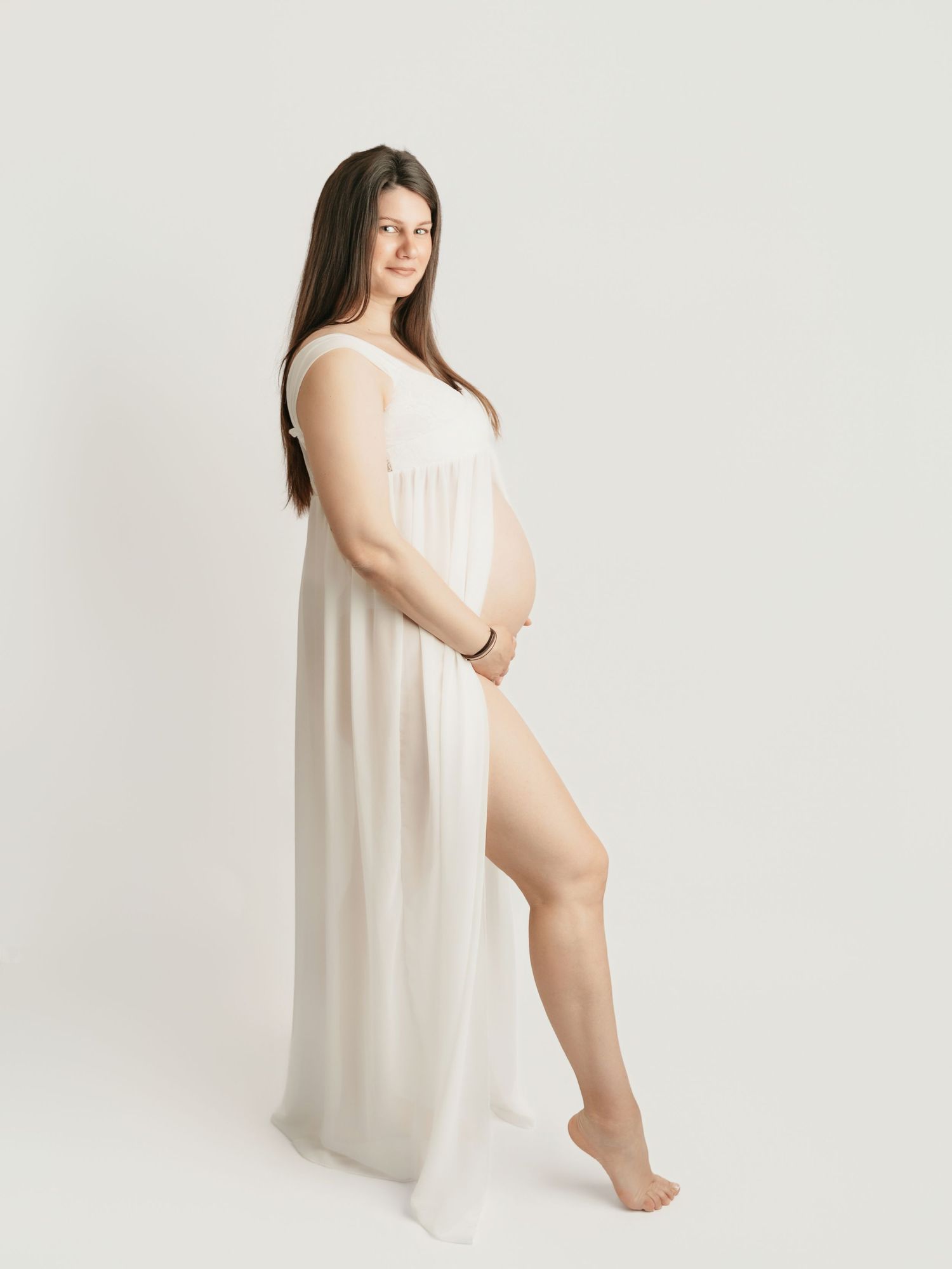 Pregnant woman in flowing white sheer gown posing sideways cradling belly in professional maternity shoot.