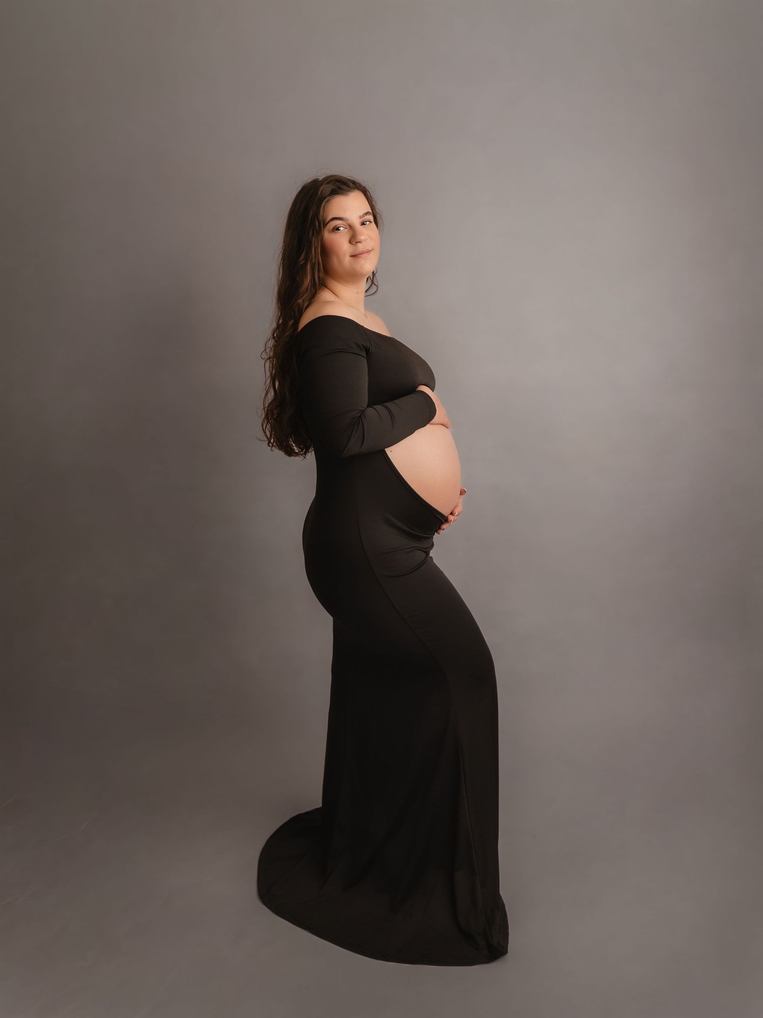 Pregnant woman in black off-shoulder fitted maternity gown posing sideways in full-length studio portrait.