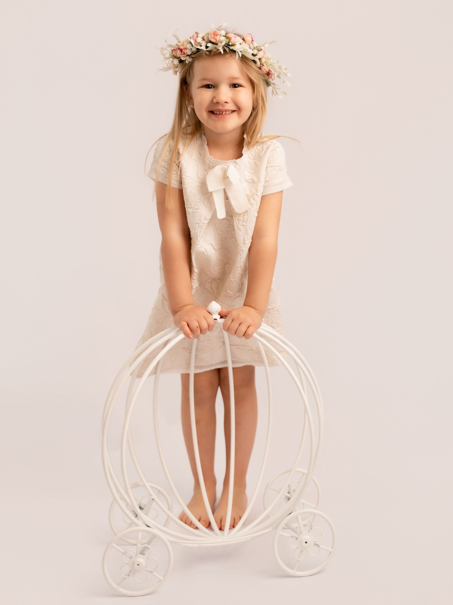 A smiling young girl in a white lace dress and floral crown poses with a decorative carriage prop.