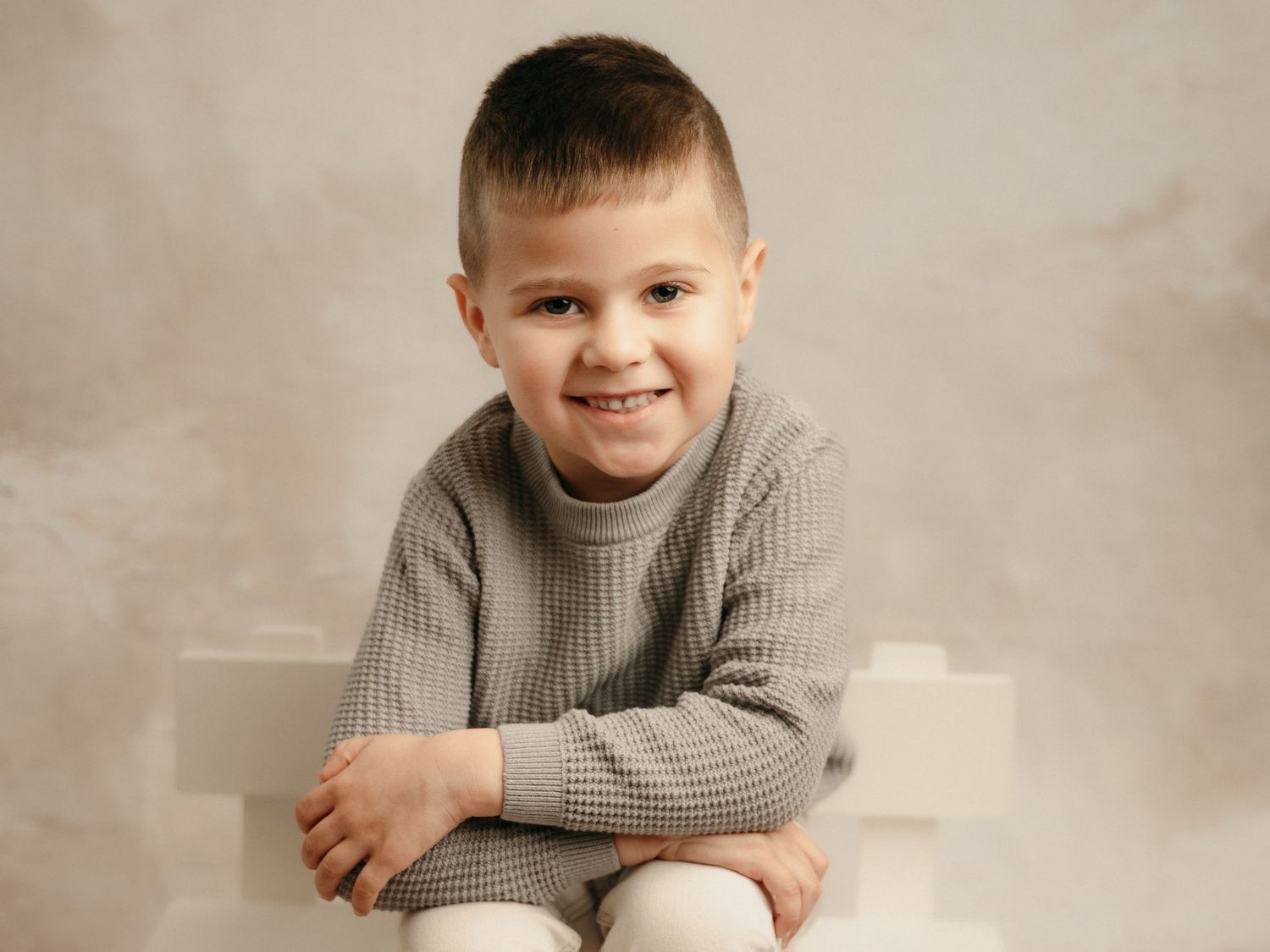 Adorable smiling young boy in gray waffle knit sweater seated on white blocks in a studio portrait.