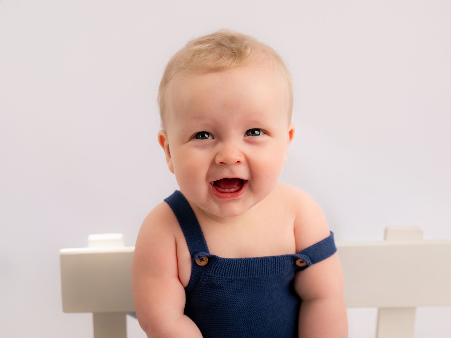 Cheerful baby in navy blue overalls sitting on a white chair, smiling with tiny teeth showing.