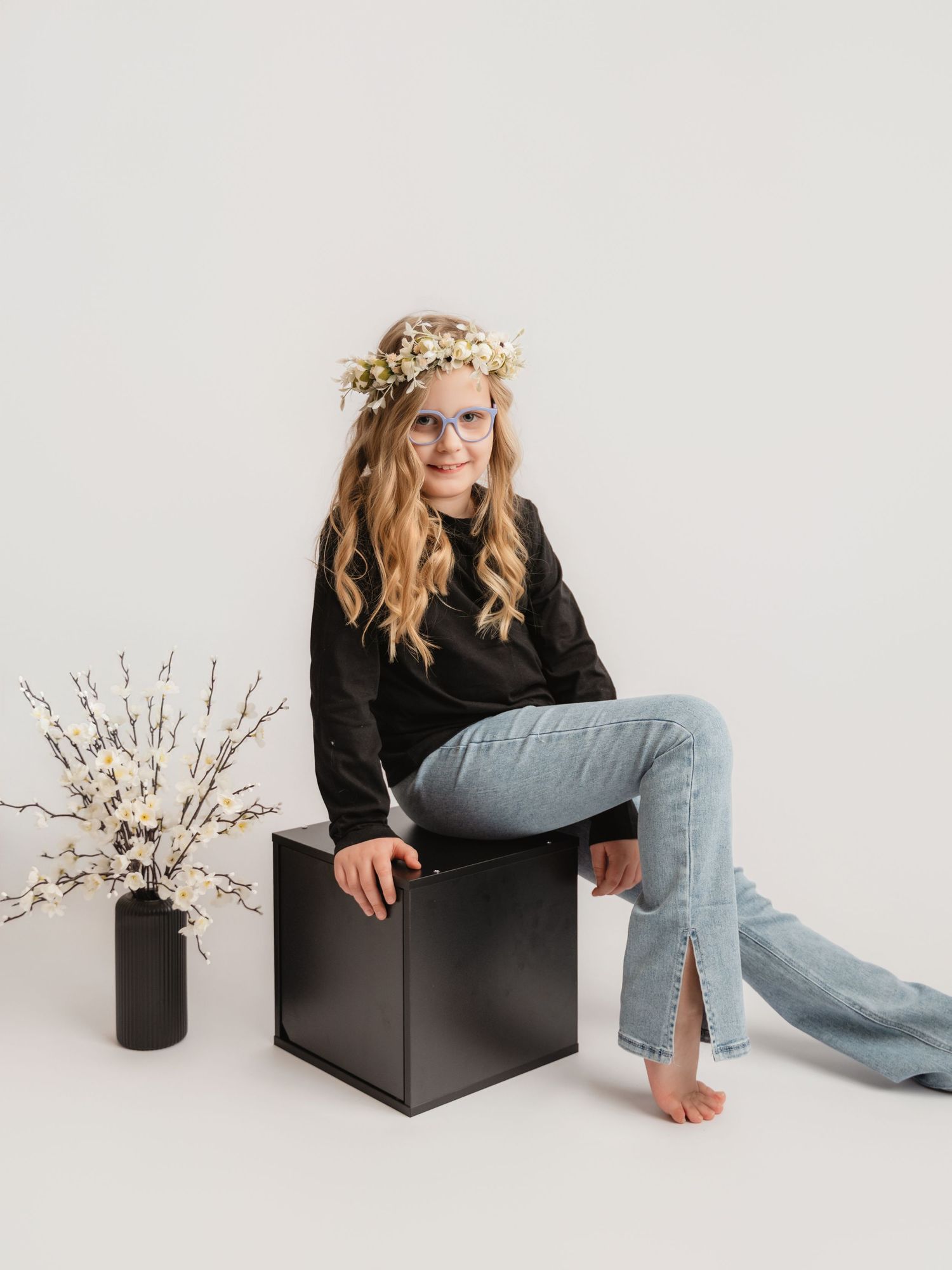 Young girl with floral crown sits on black cube in black top and jeans beside white blossoms.