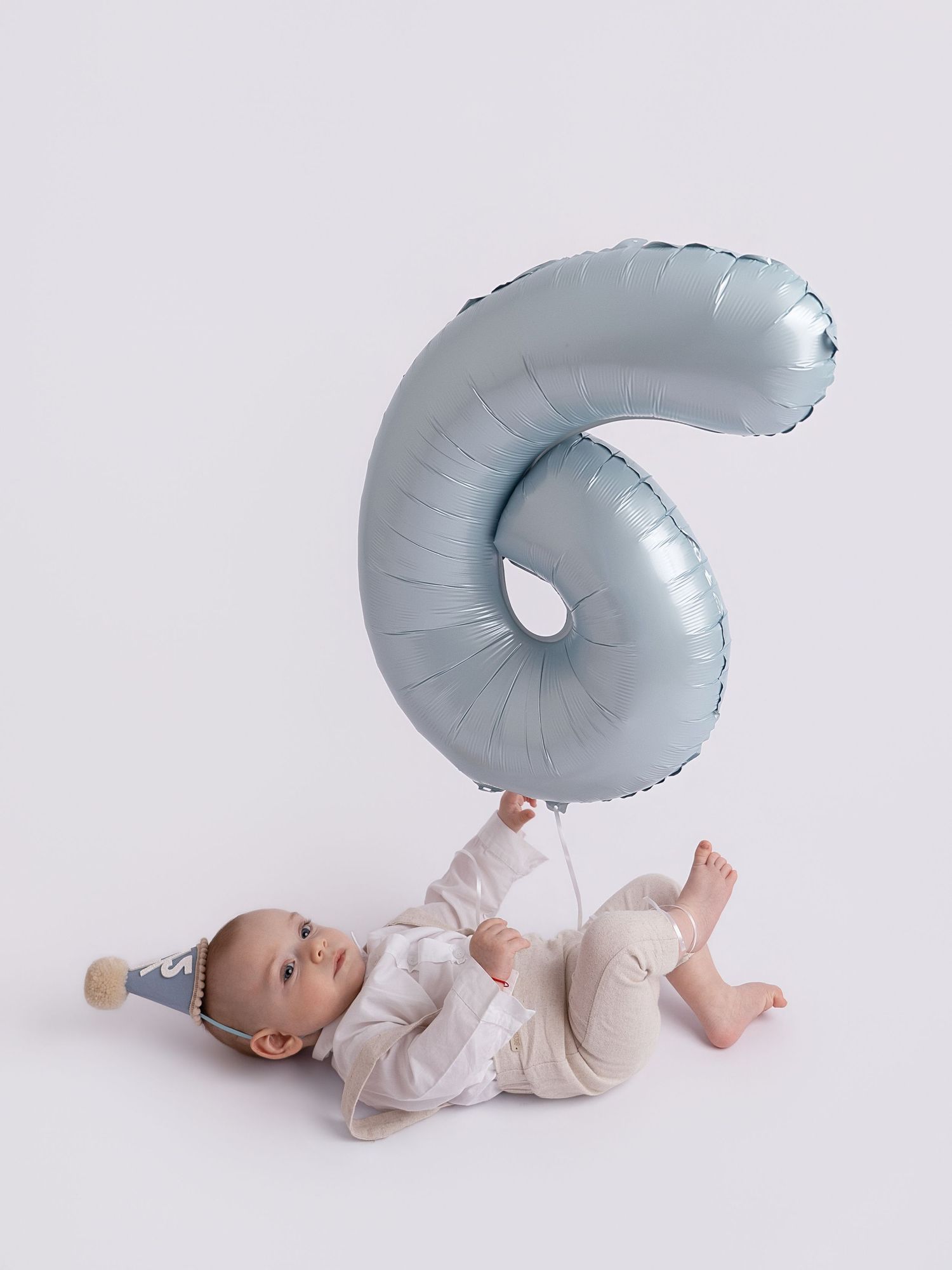 Baby lying on back holding a light blue number 6 foil balloon, wearing a party hat, celebrating 6 months old.