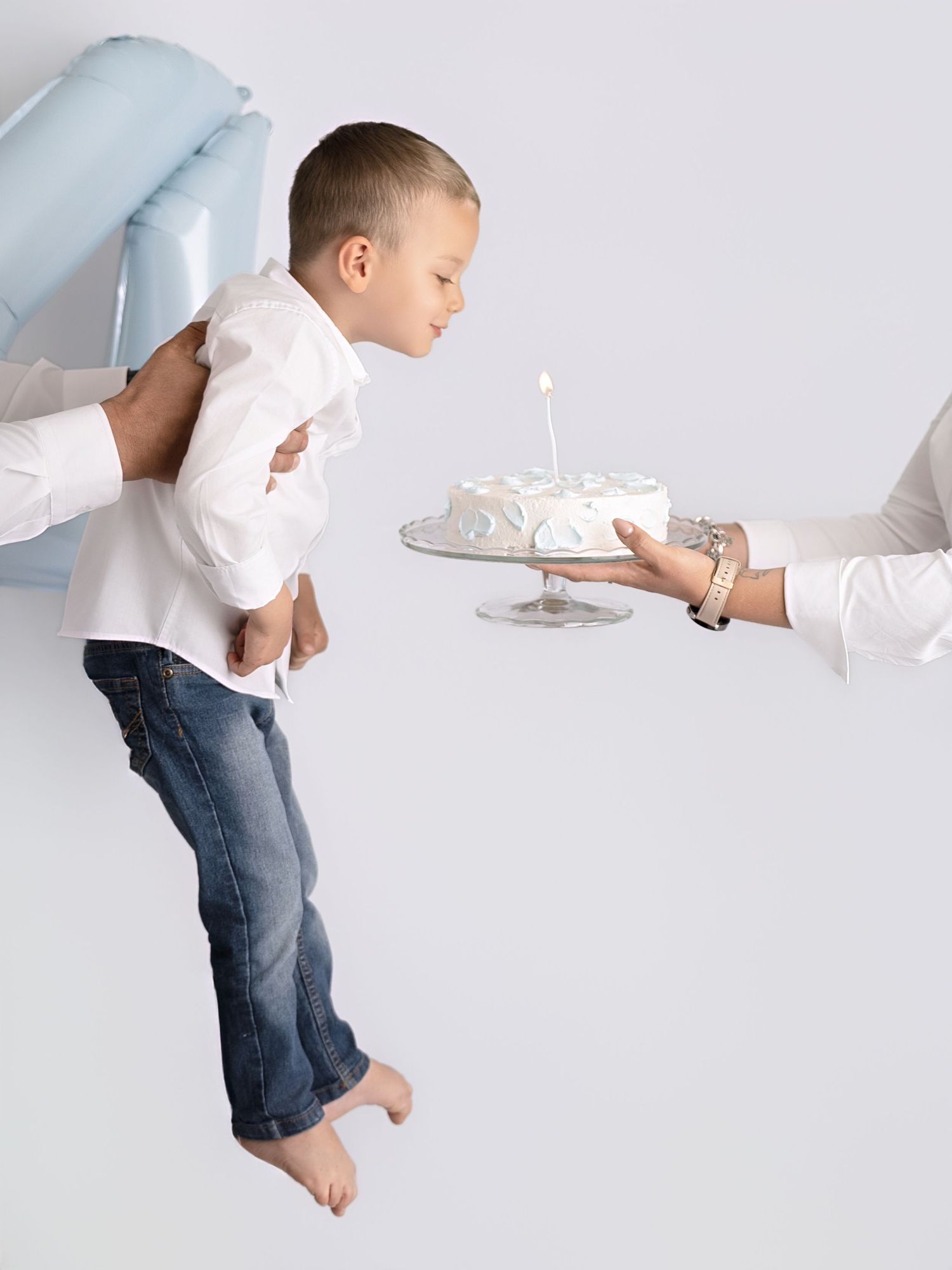 Boy in white shirt blows out candle on white birthday cake held by adult hands in studio photo.