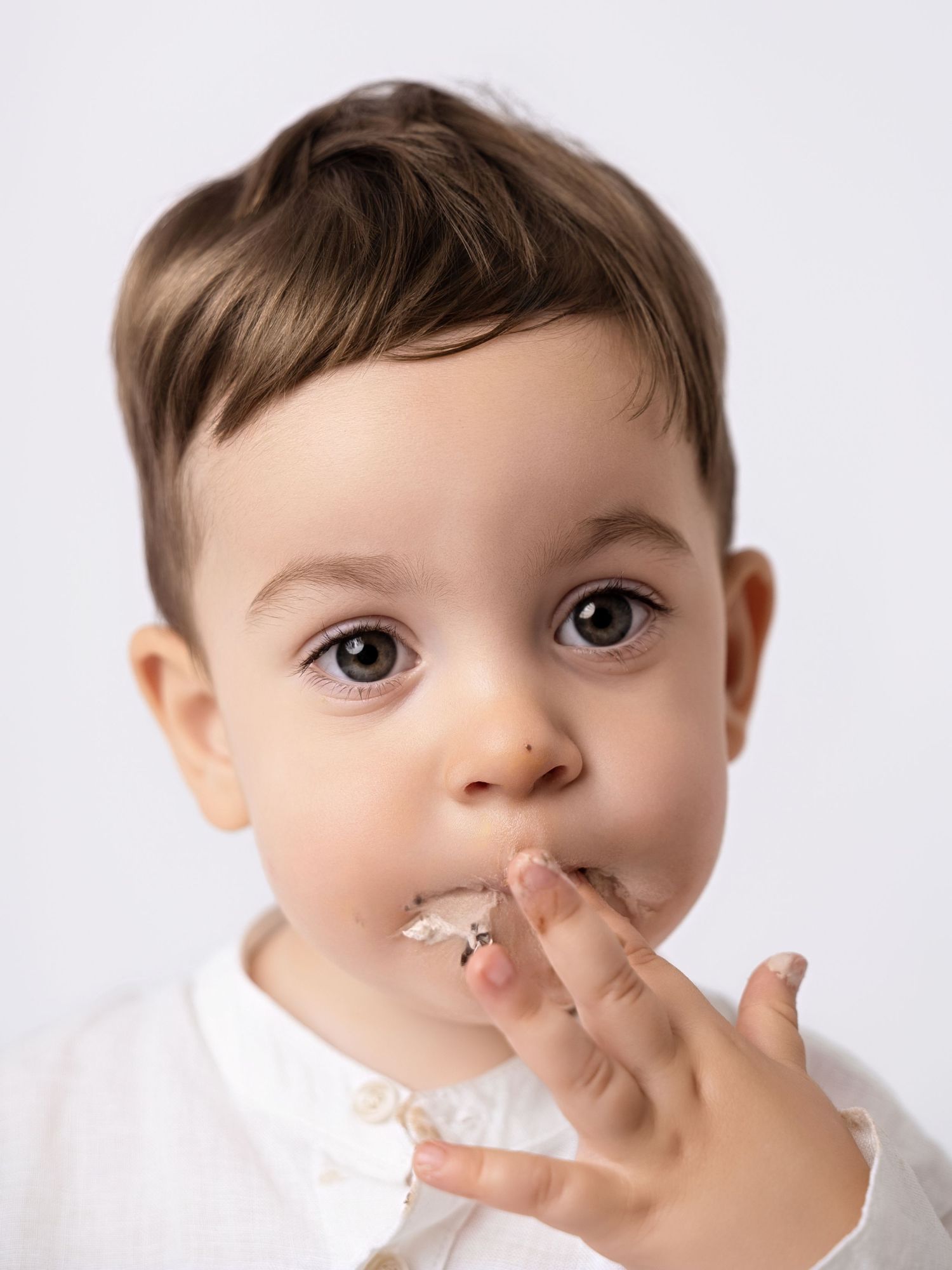 Cute toddler boy with brown eyes licking frosting off his finger against a white background.