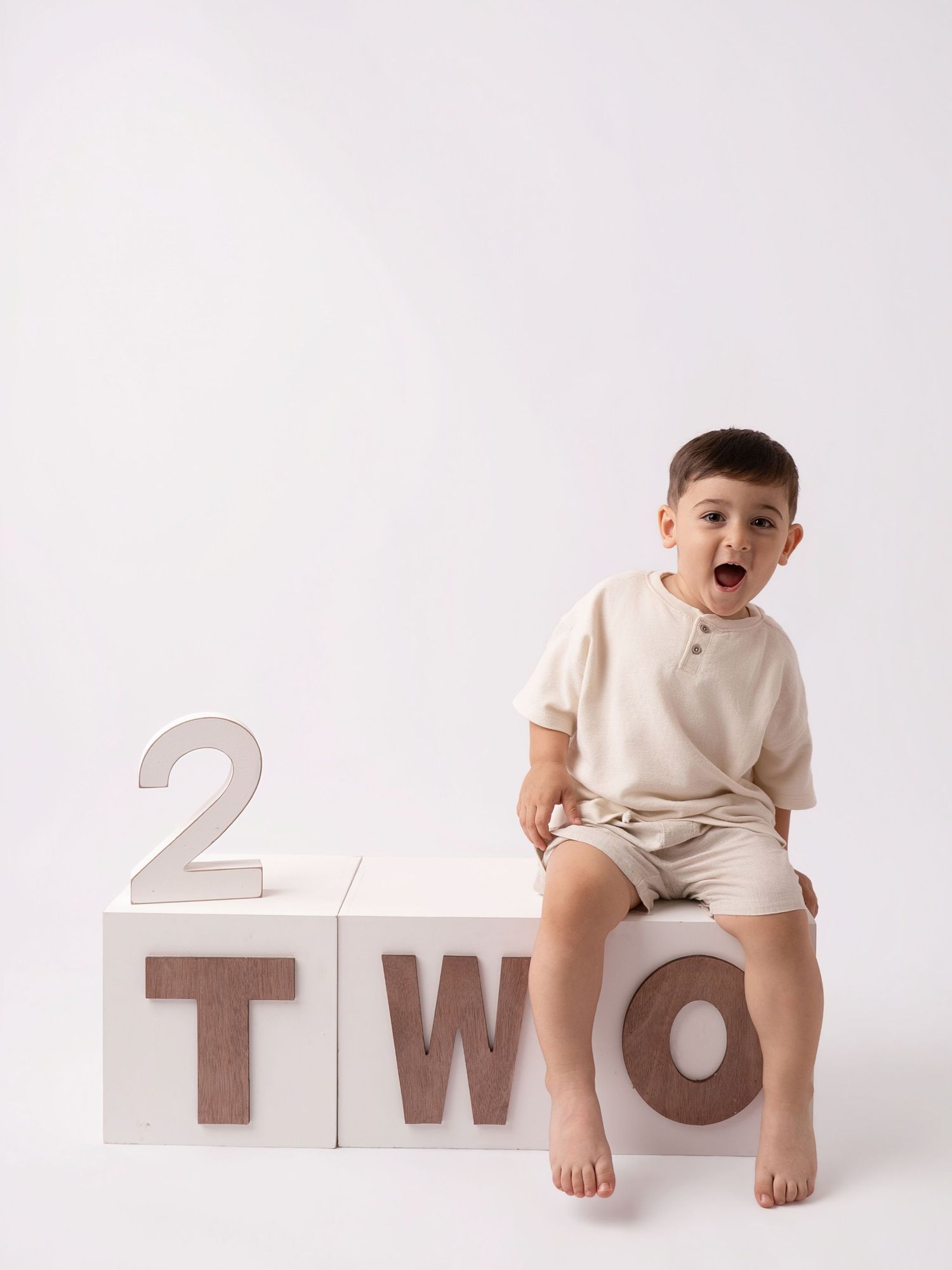 Toddler boy sitting on white blocks spelling TWO for his 2nd birthday photo session.