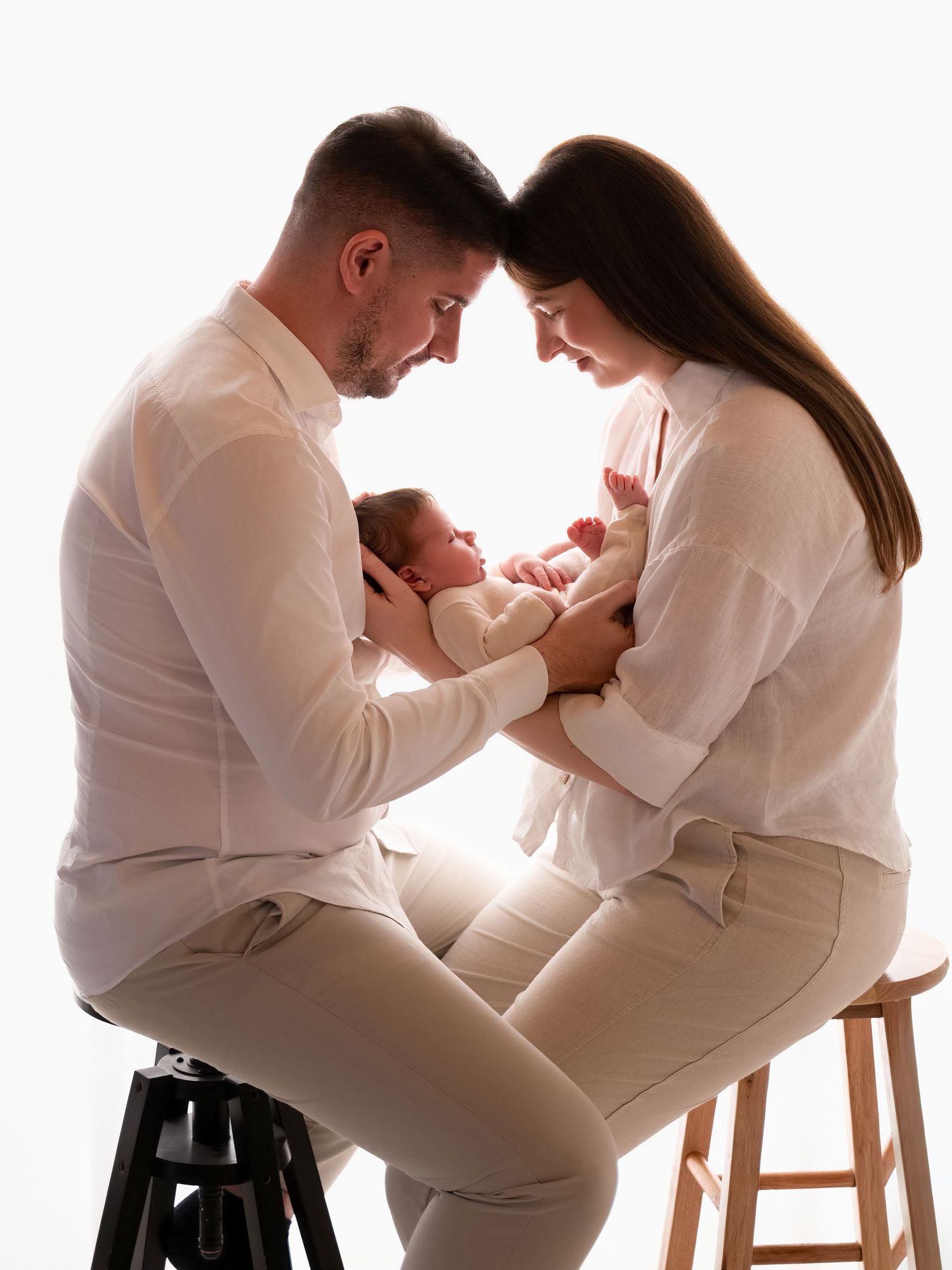Parents in white outfits tenderly hold their newborn baby together, foreheads touching in a bright studio portrait.