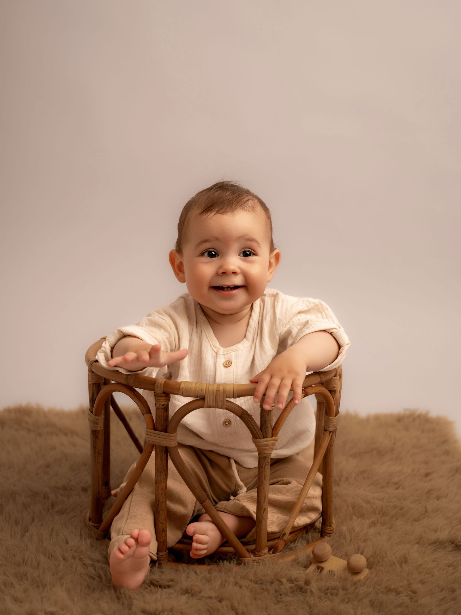 Smiling baby in cream outfit sitting inside a rattan basket on a fluffy brown rug during a studio photo session.
