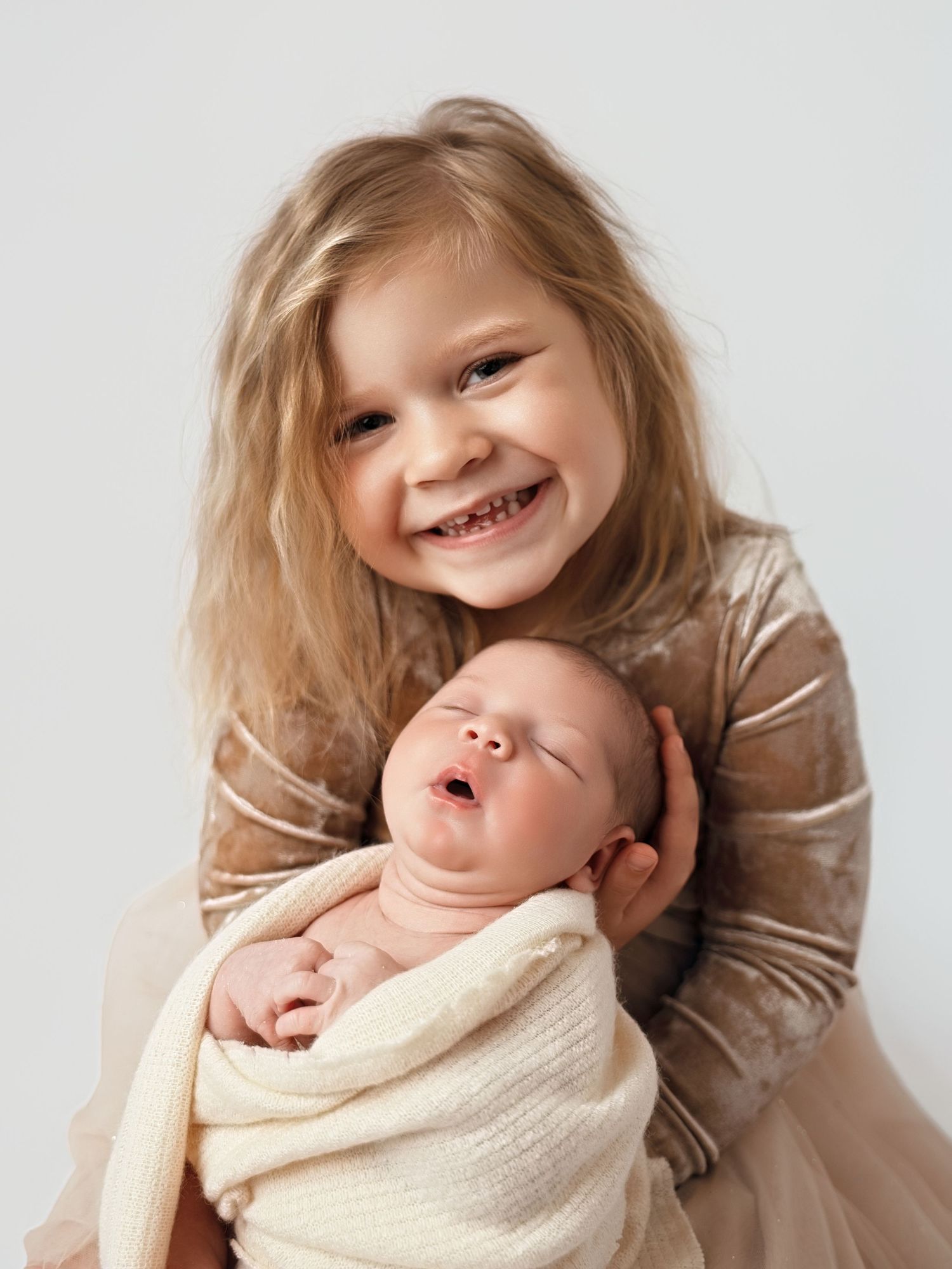 Young girl smiling brightly while holding a swaddled newborn baby sibling in a soft, neutral-toned portrait.