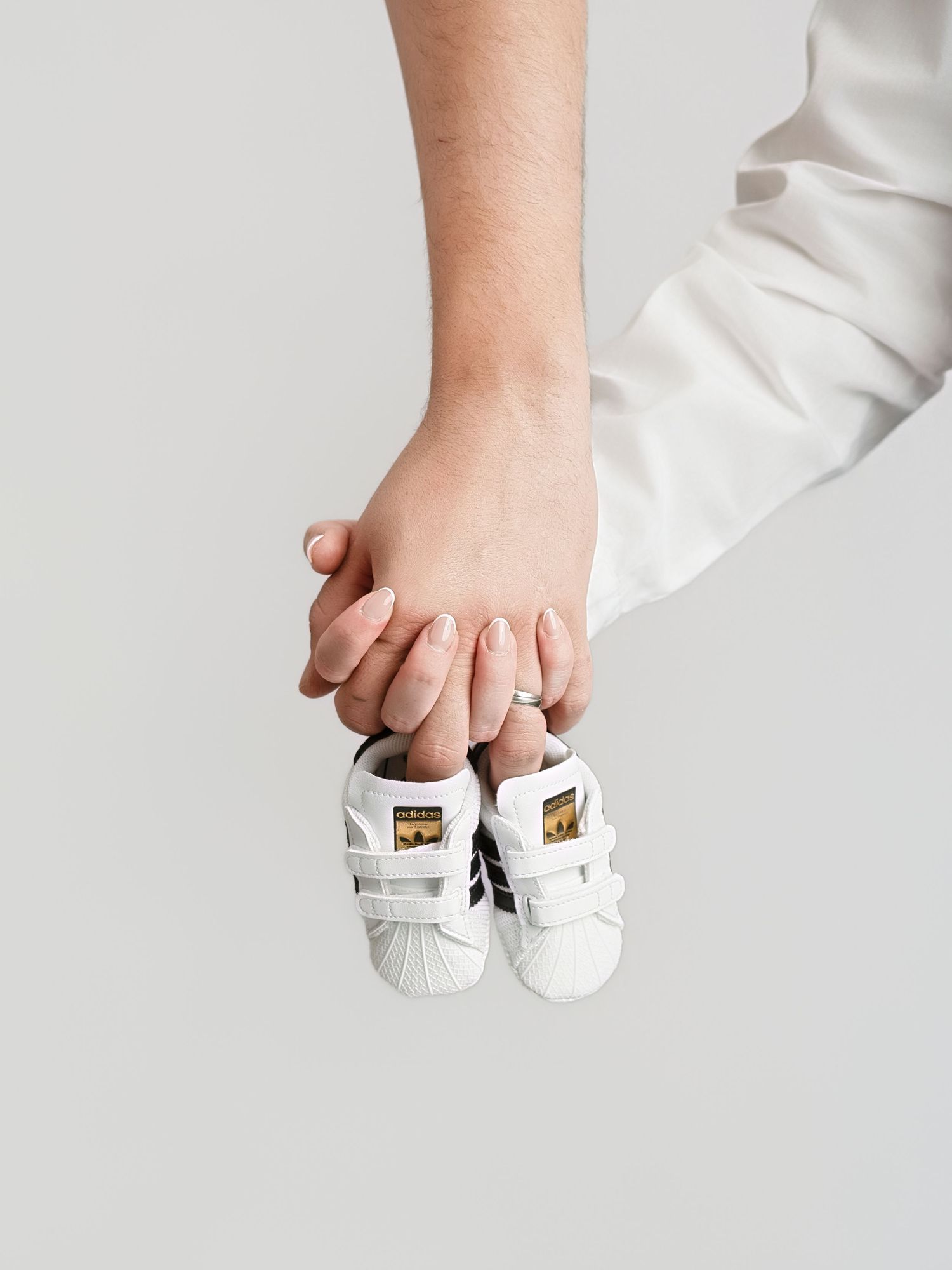 Two hands holding tiny white Adidas Superstar baby sneakers with black stripes against a light gray background.