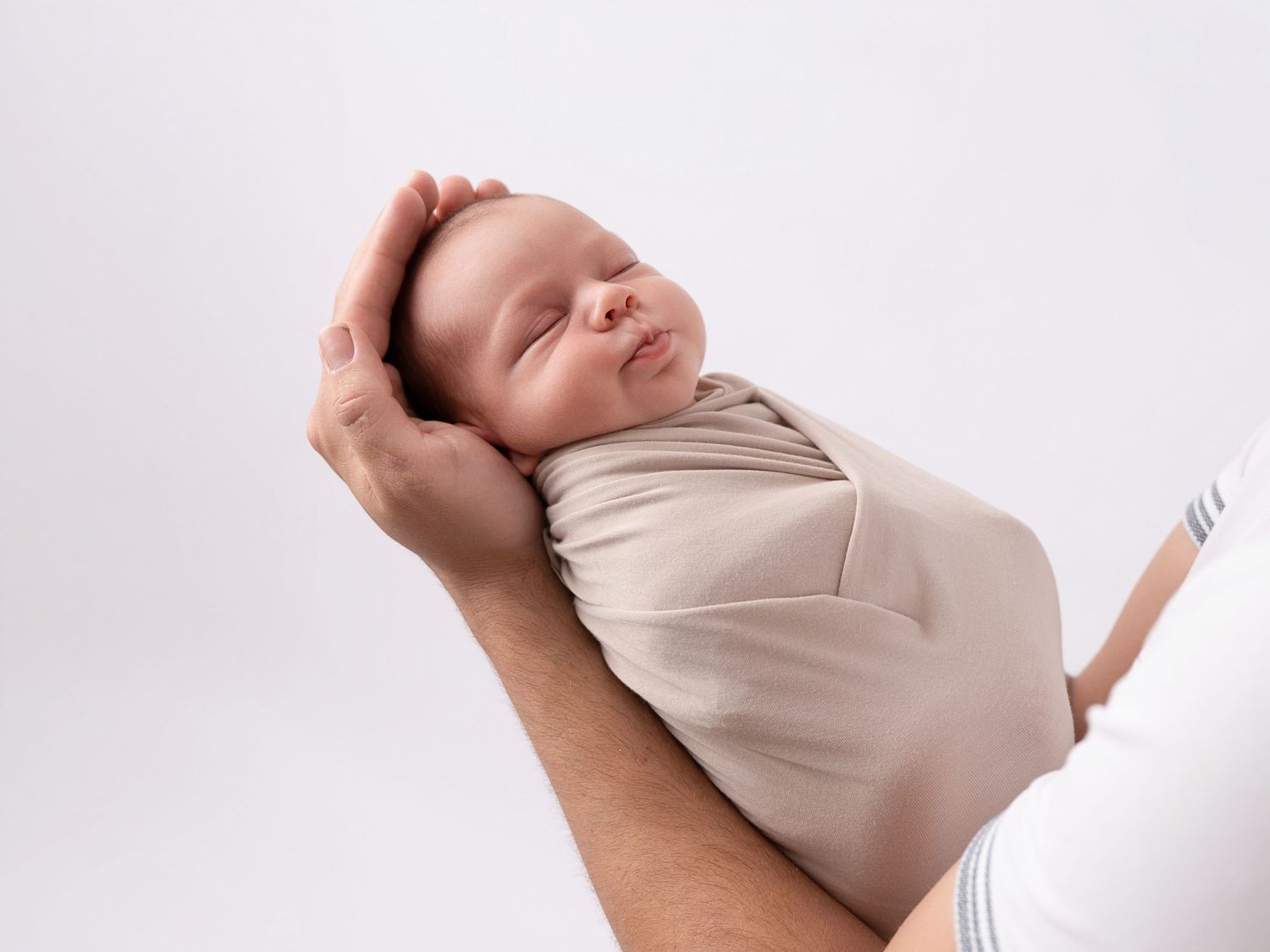 Newborn baby swaddled in beige wrap sleeping peacefully in adult hands against white background.