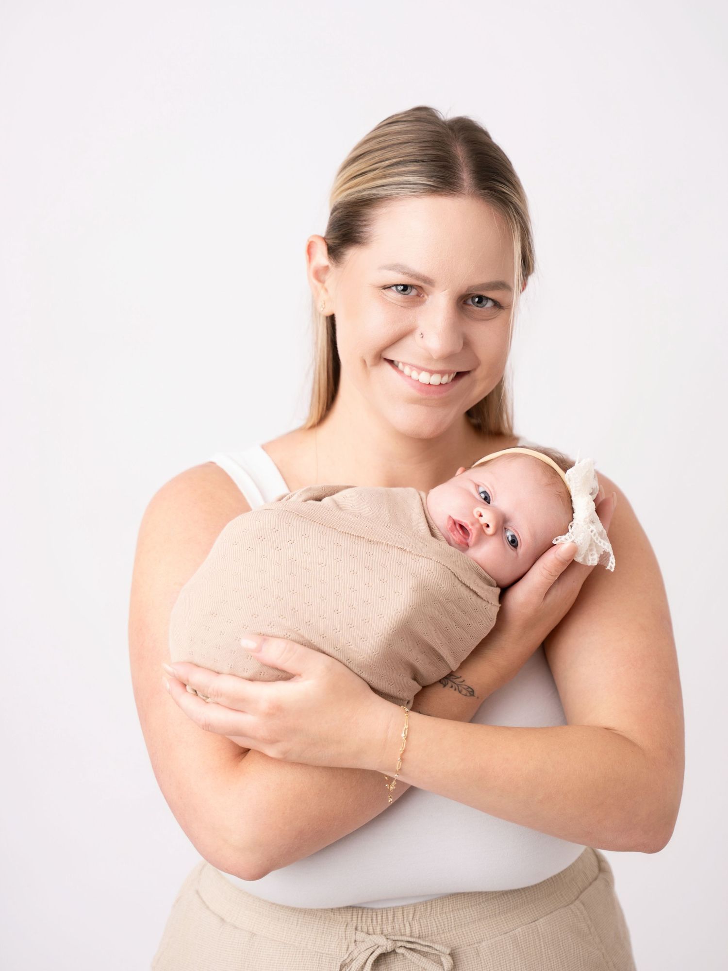 Smiling mother holding her swaddled newborn baby wearing a white headband against a white background.
