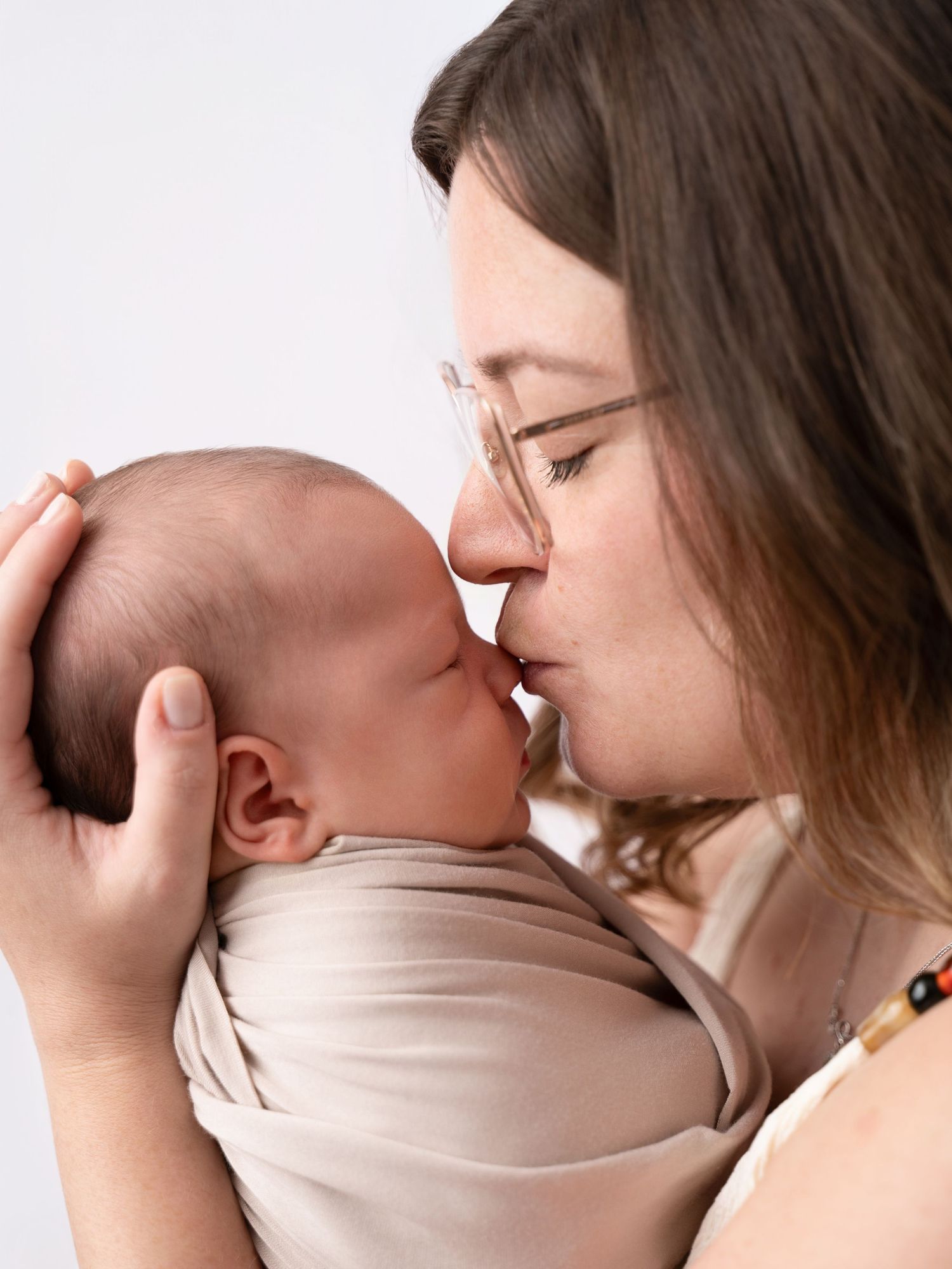 A mother gently kisses her sleeping newborn baby wrapped in a beige swaddle against a white background.