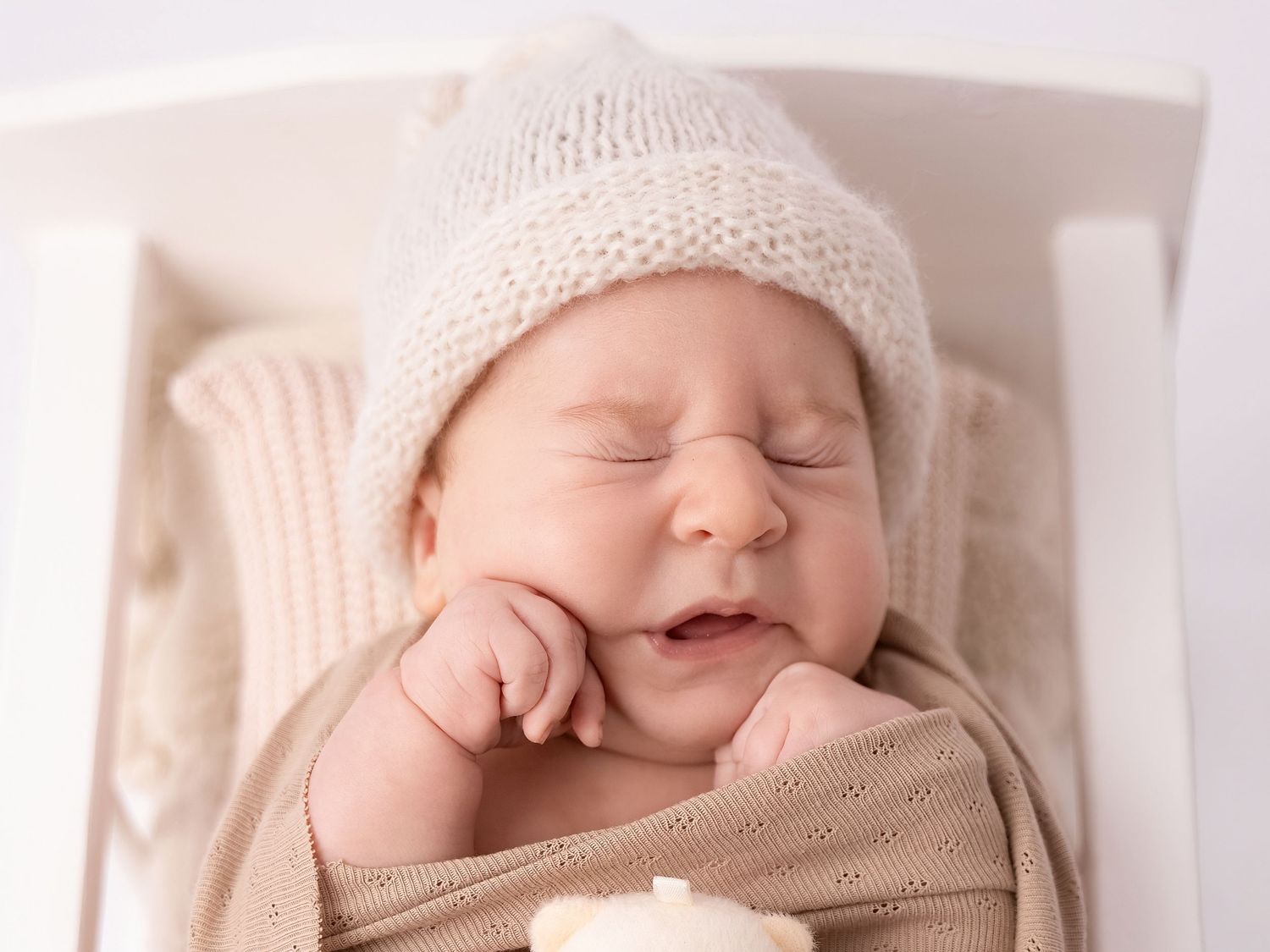 Peaceful newborn baby sleeping in white crib, wearing cozy knit beige hat, wrapped in soft neutral swaddle blanket.