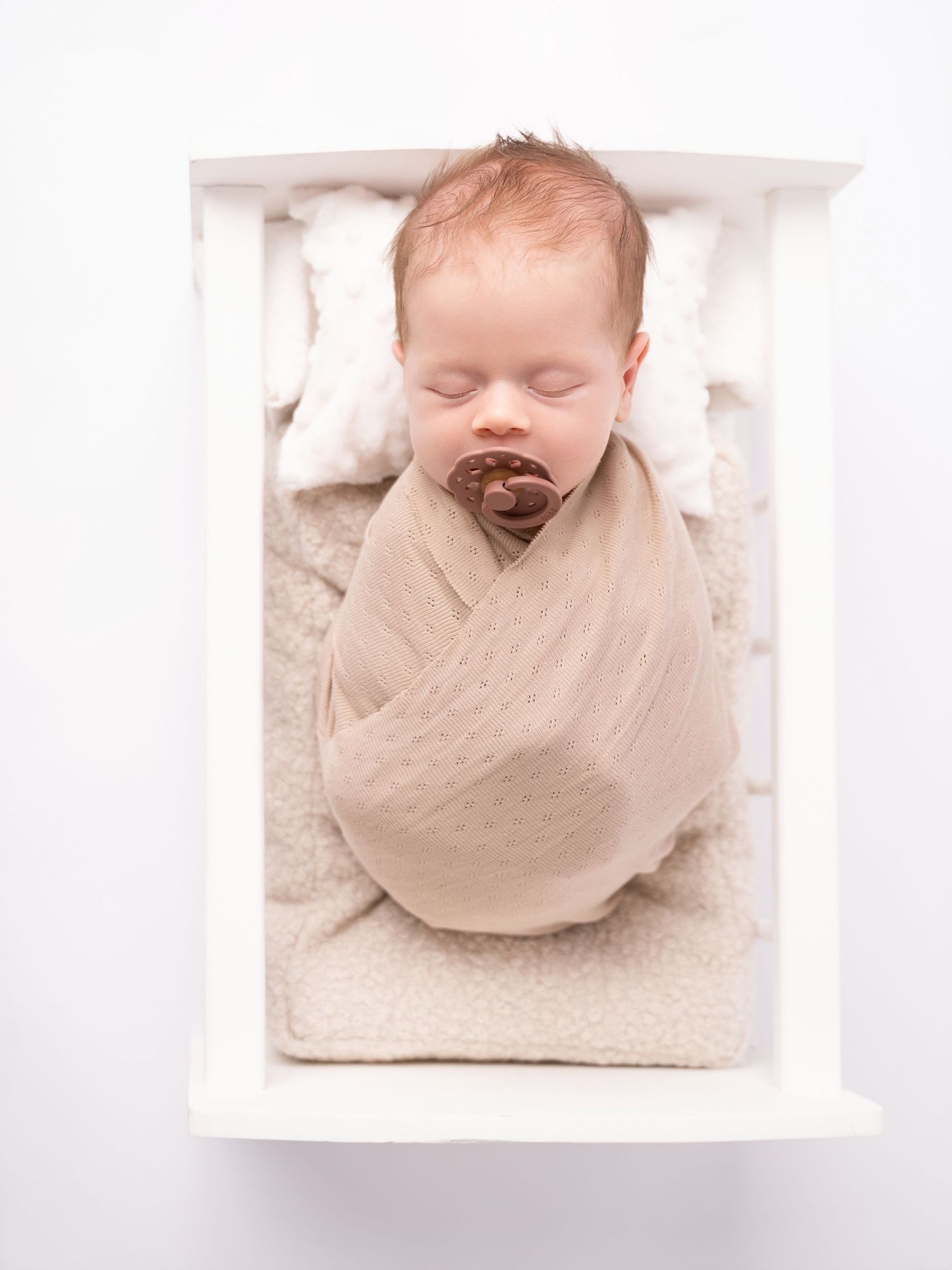 Sleeping newborn baby swaddled in beige wrap with brown pacifier lying in white wooden crib on fluffy white bedding.