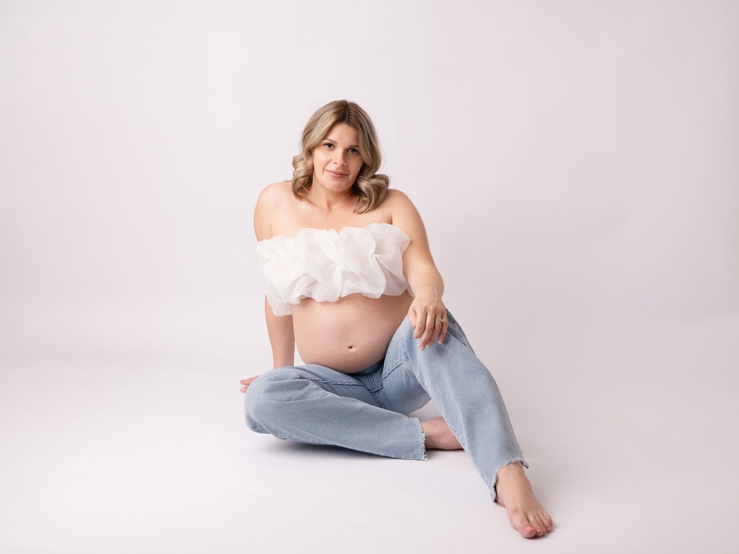 Pregnant woman in white ruffled crop top and light blue jeans sitting on floor against white background.