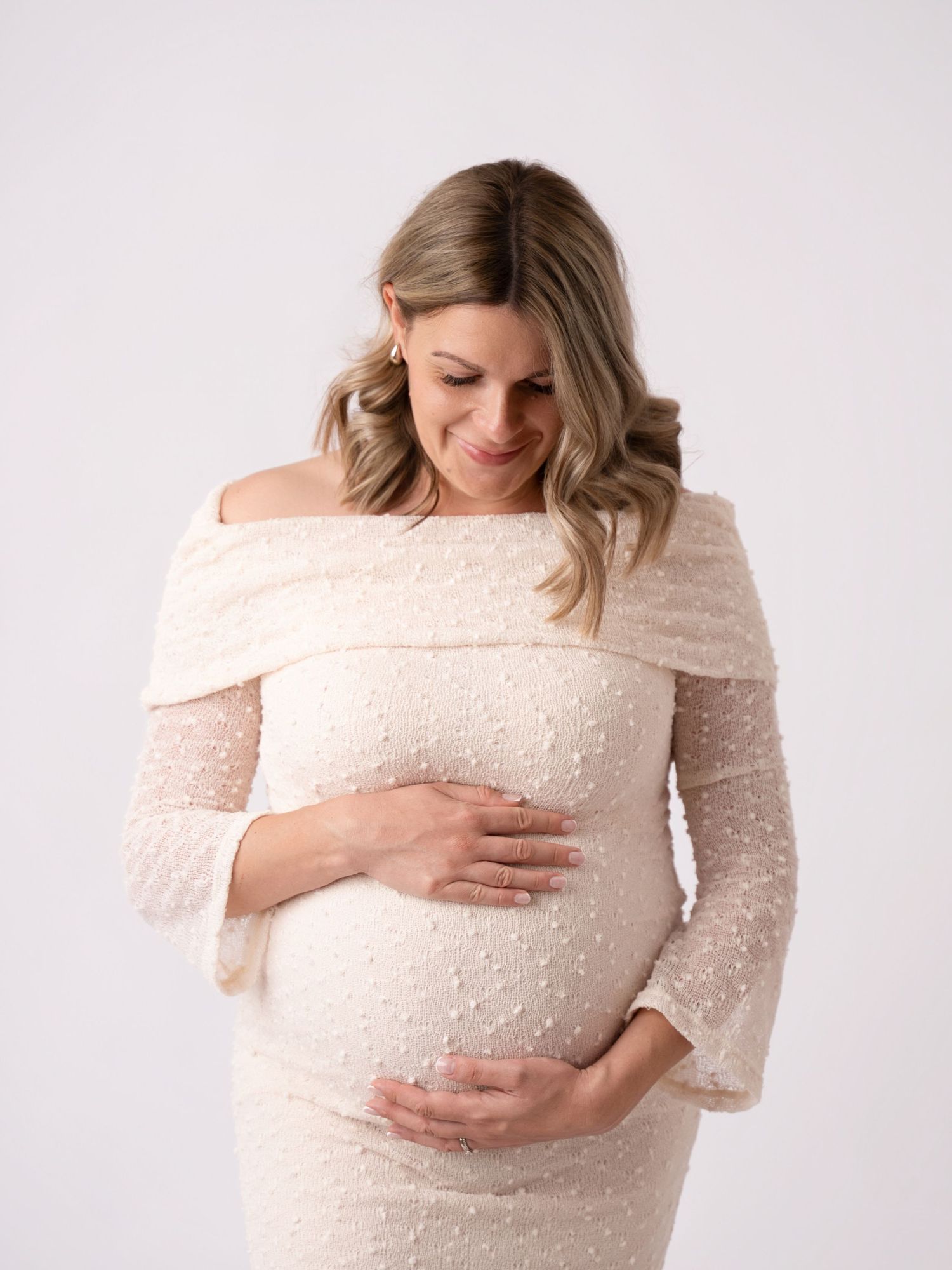 Pregnant woman in elegant off-shoulder cream lace dress cradling her baby bump, smiling softly in a studio setting.