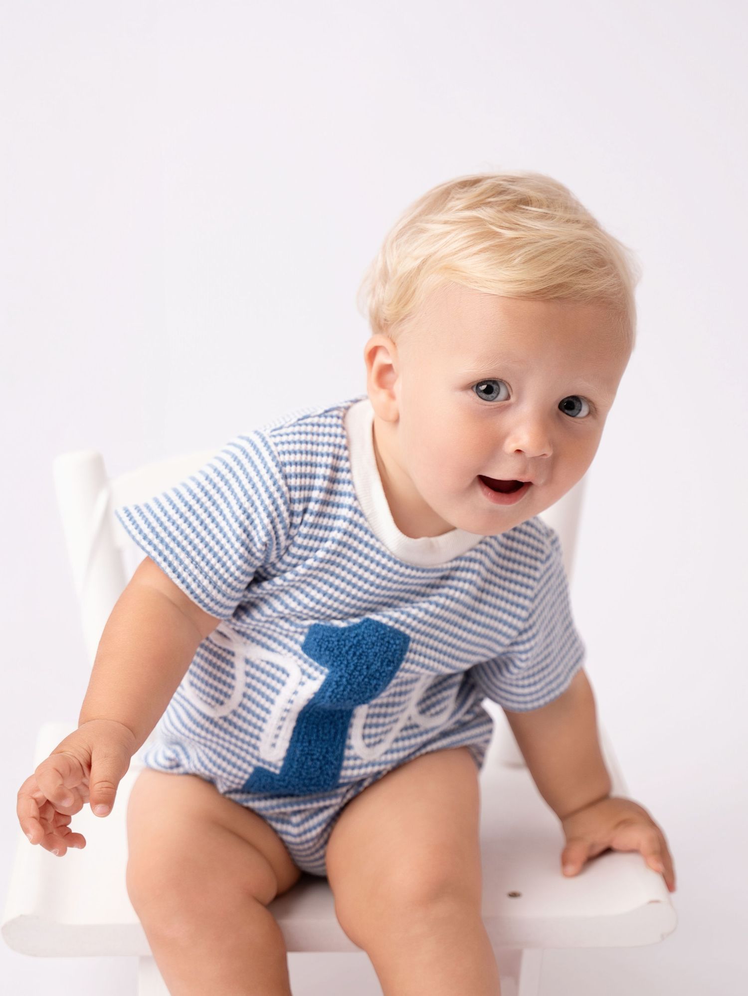 Smiling baby boy in blue striped seersucker romper with number one applique sitting on white chair.