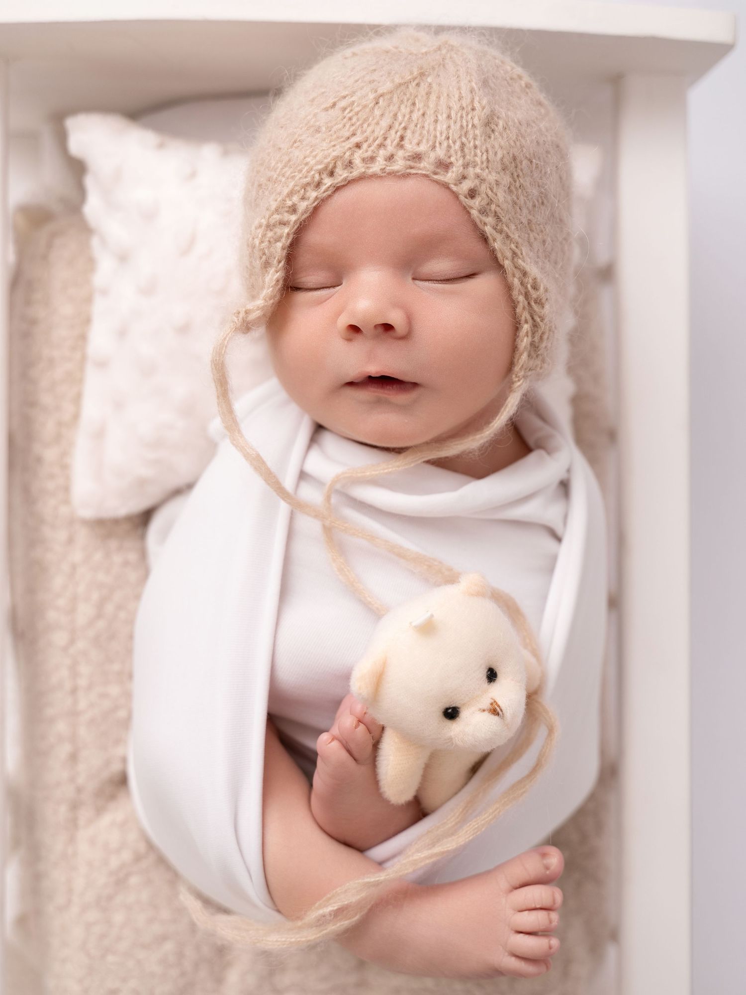 Sleeping newborn baby swaddled in white wrap wearing a beige knit bonnet holding a small stuffed animal toy.