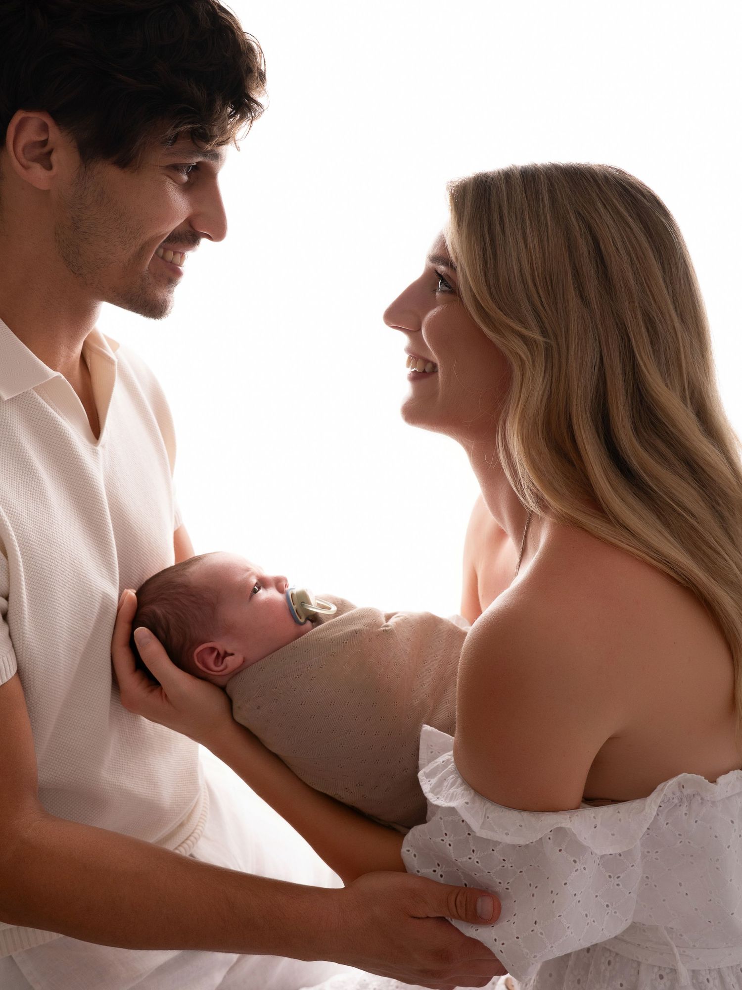 A couple smiles at each other while tenderly holding their sleeping newborn baby wrapped in a soft blanket.