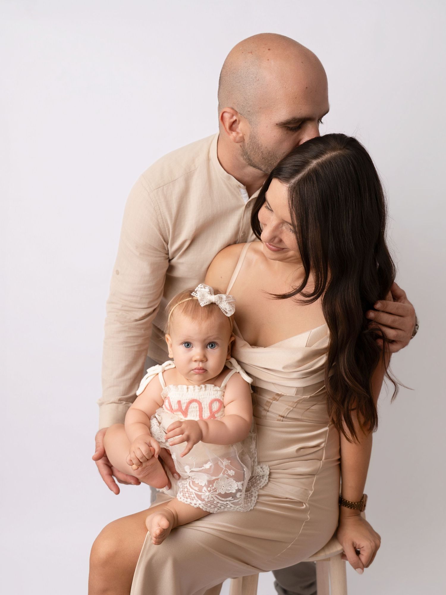 A bald man kisses a brunette woman holding a baby girl in a white outfit during a family portrait session.