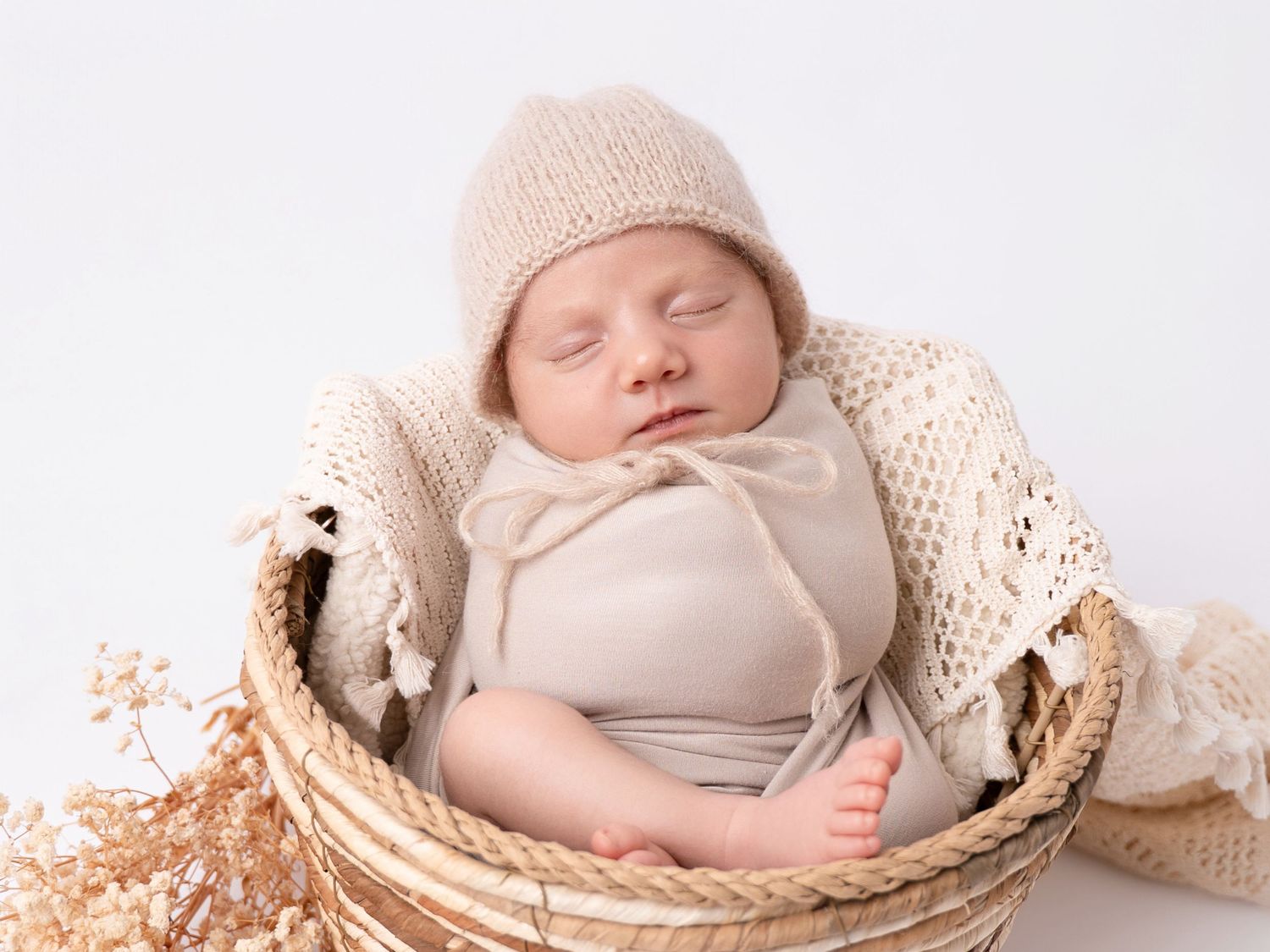 Sleeping newborn baby in beige knit hat and wrap, nestled in a wicker basket with cream blanket.
