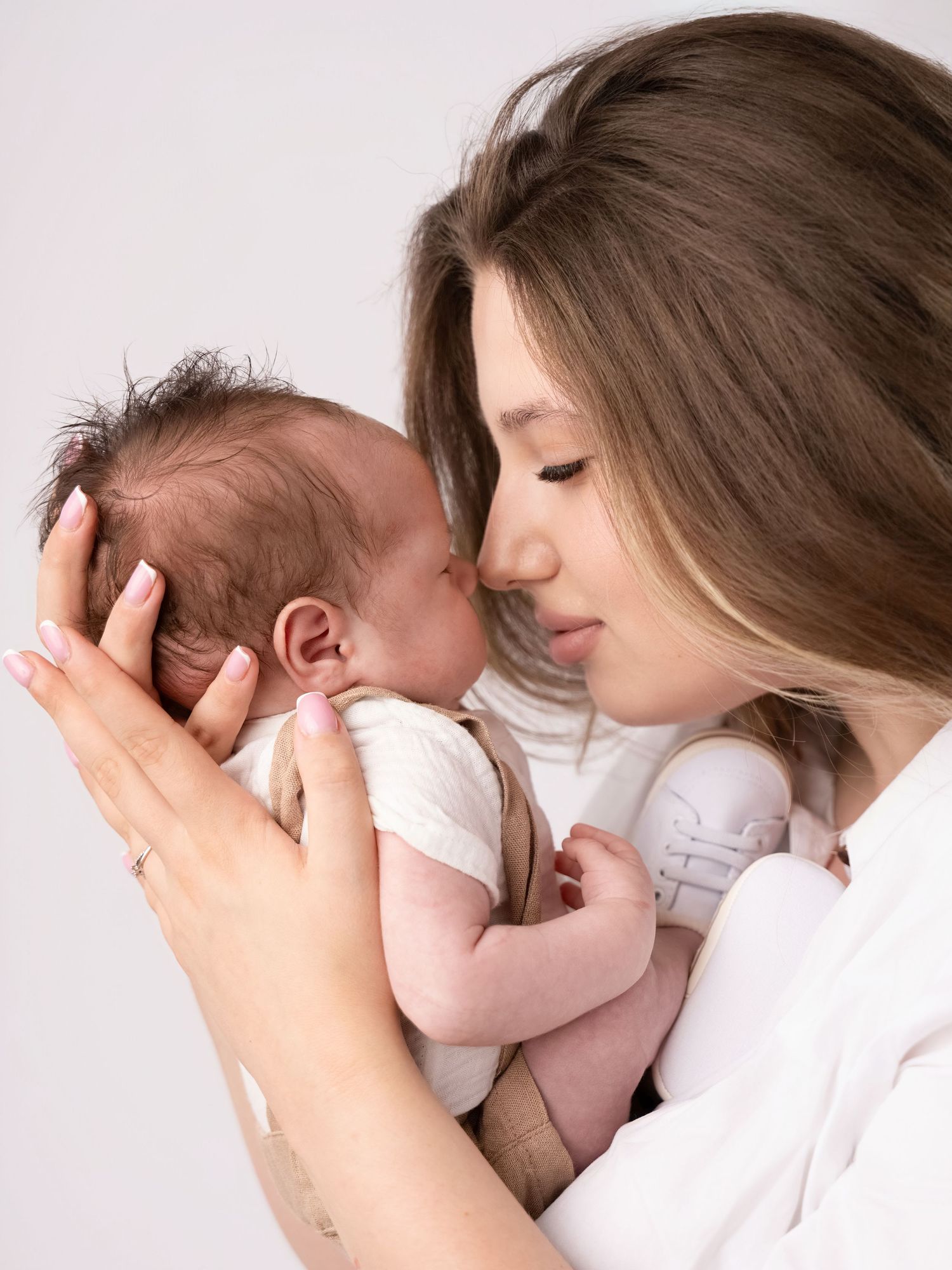 A young mother tenderly kisses her newborn baby while cradling it gently in her hands against a soft background.