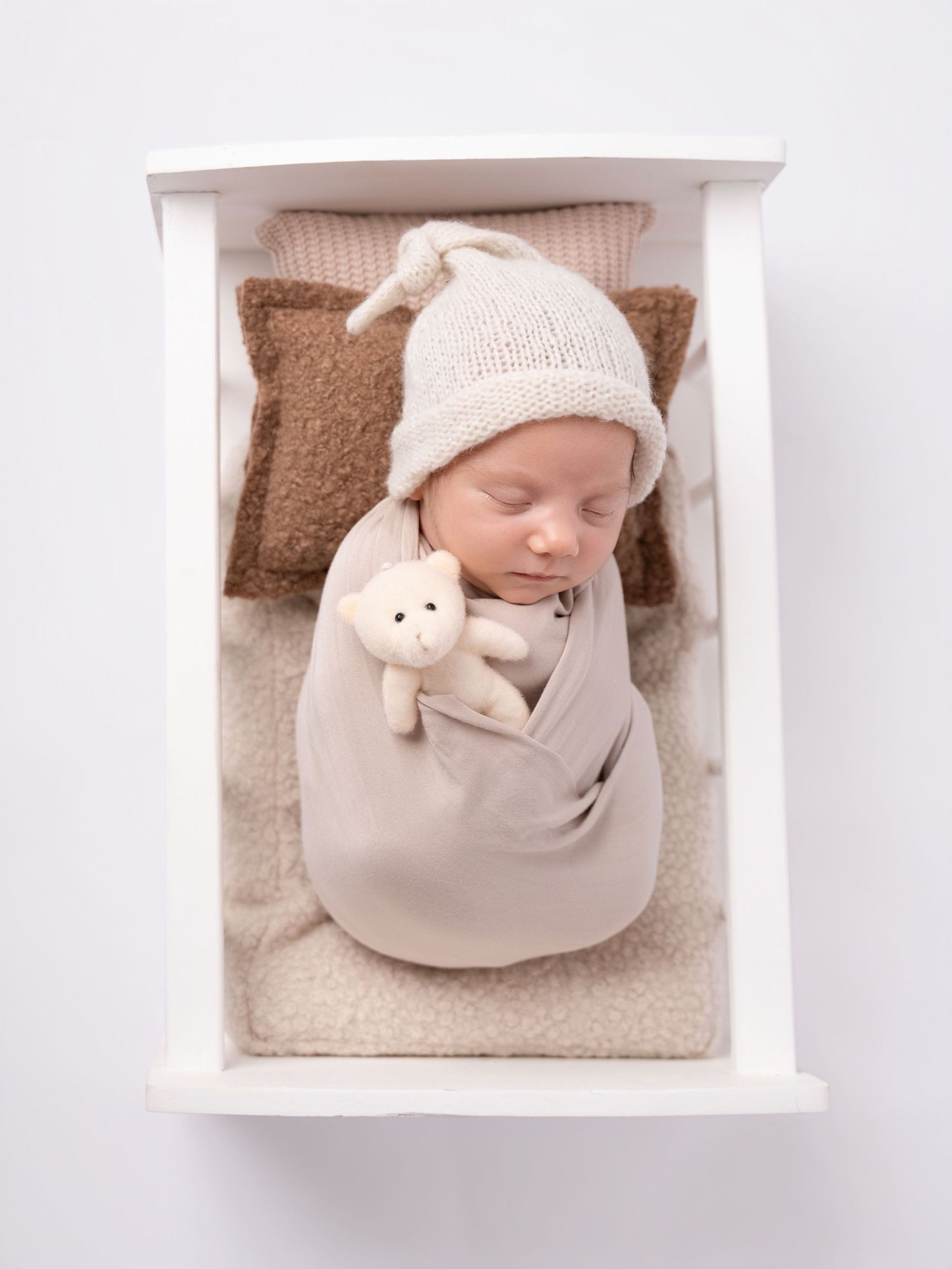 Swaddled newborn baby sleeping in a white wooden box with knit hat and tiny teddy bear.
