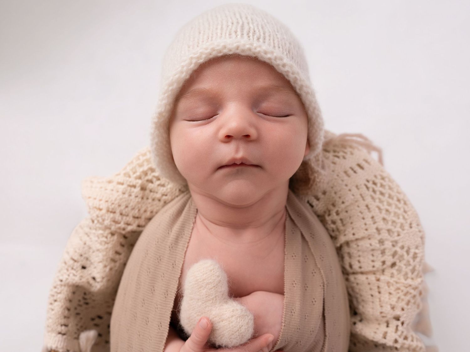 Peaceful sleeping newborn baby wearing a cream knit bonnet, wrapped in a soft beige blanket, holding a tiny stuffed toy.