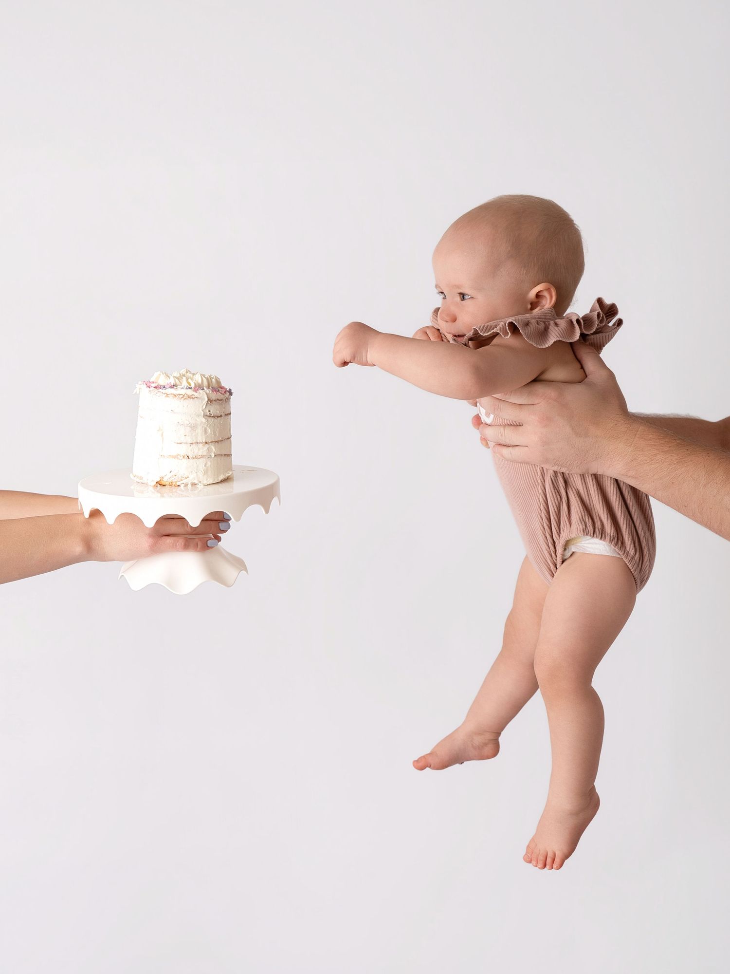 A baby in a pink romper reaches toward a small white cake on a stand, held by an adult hand, against a white background.
