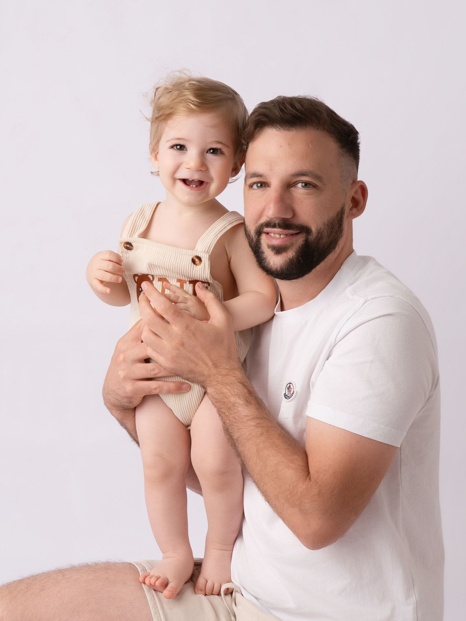A smiling man in a white t-shirt holds a happy toddler in a beige romper against a light background.