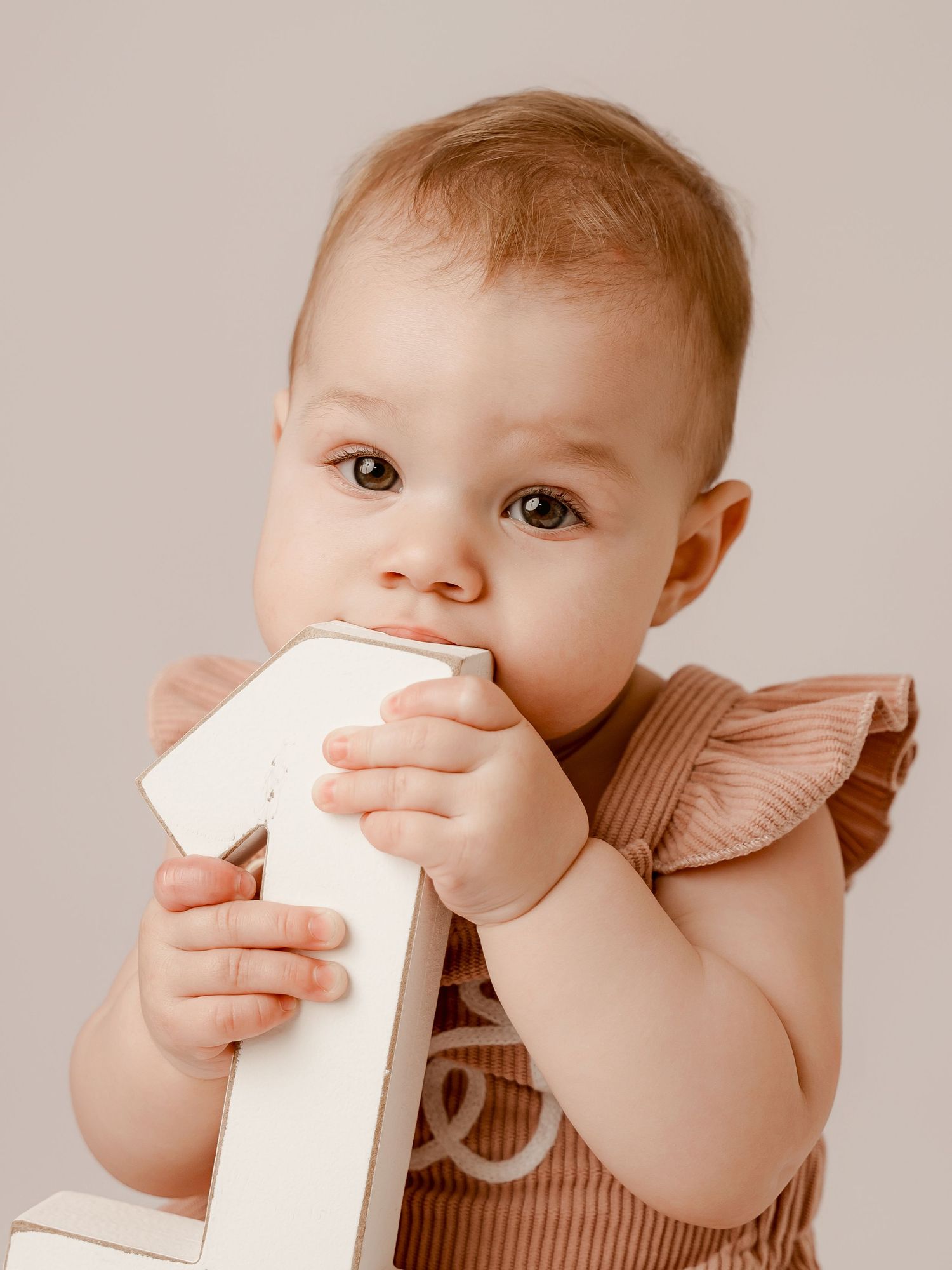 A cute baby in a brown ruffle romper chews on a white number one block, celebrating their first birthday.