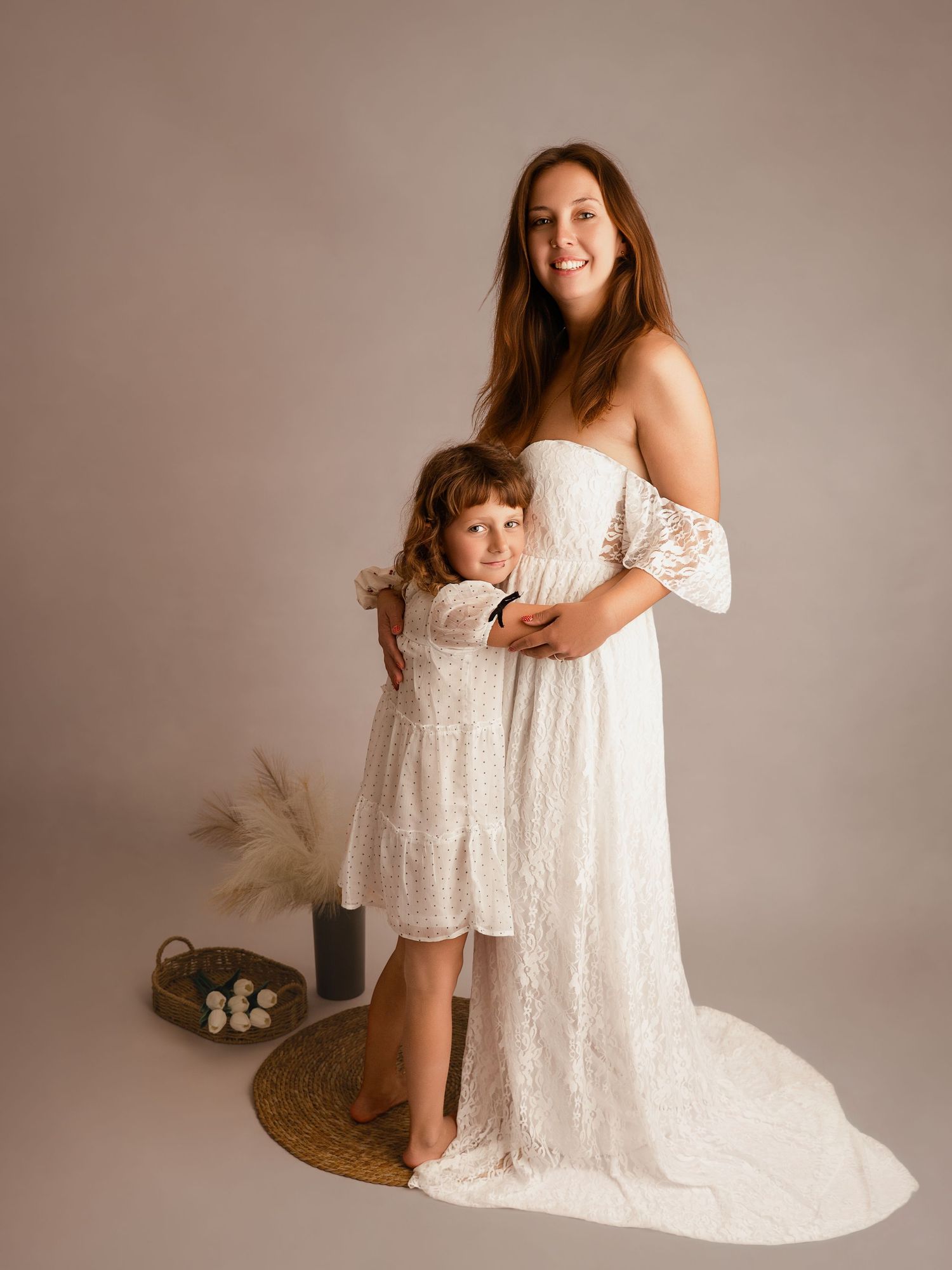 Mother in white off-shoulder gown and daughter in matching dress share a tender studio portrait.