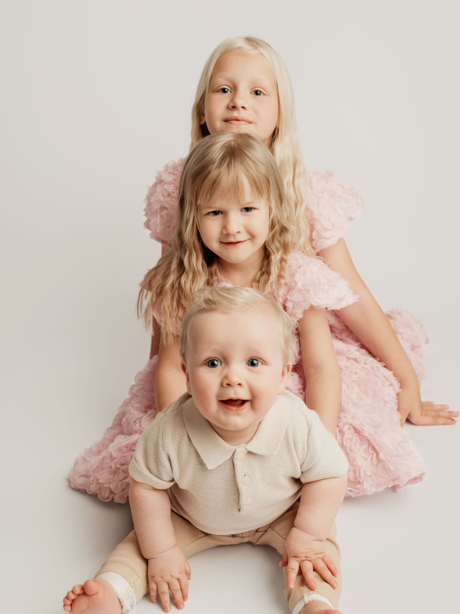 Three siblings pose together in a studio, two girls in pink dresses with a smiling baby boy in front.