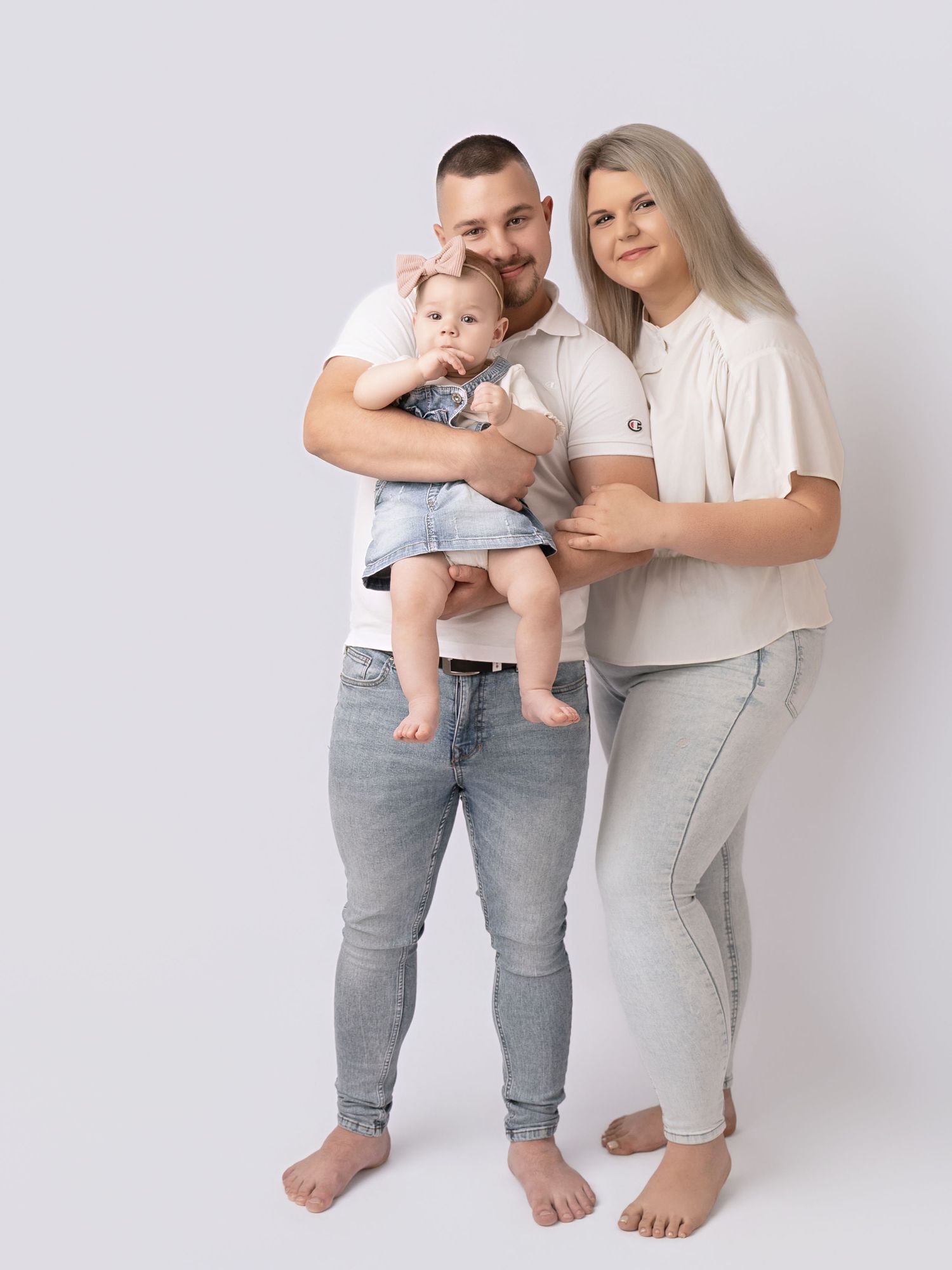 A young family poses in matching white tops and jeans, dad holding baby girl in a bright studio.