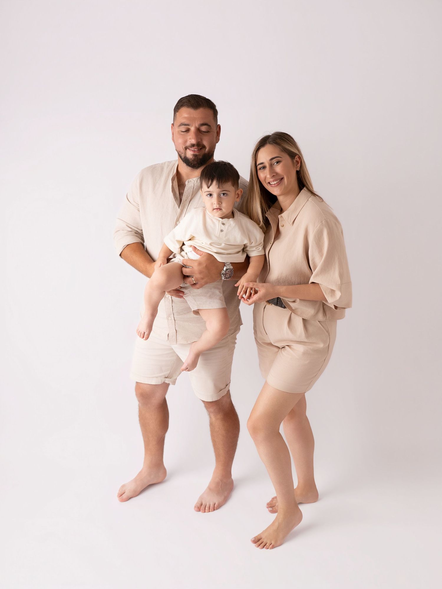 Family of three in matching beige linen outfits posing together, parents holding baby in bright studio.