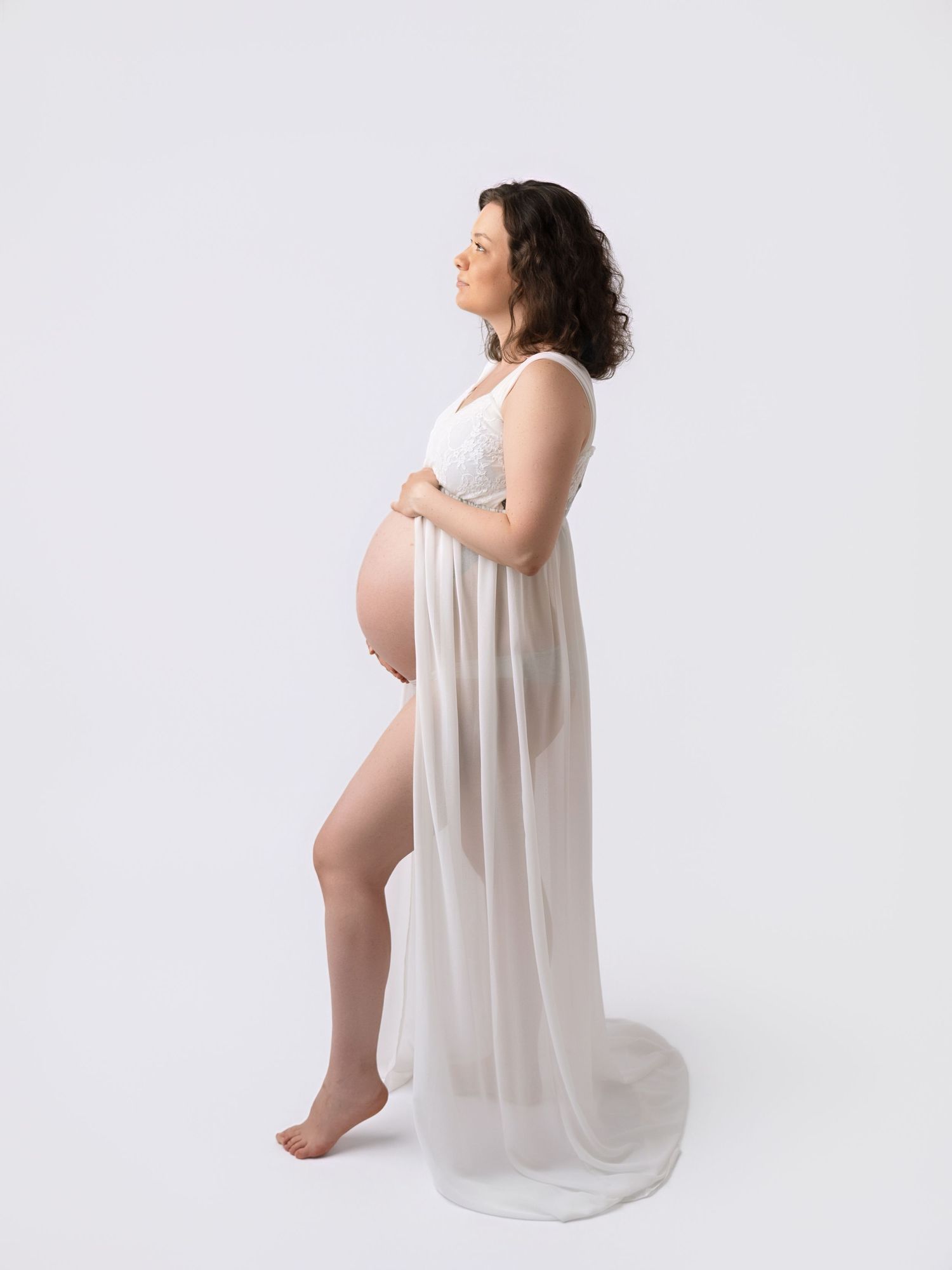 Pregnant woman in white sheer maternity gown holding baby bump, side profile studio shot on white background.
