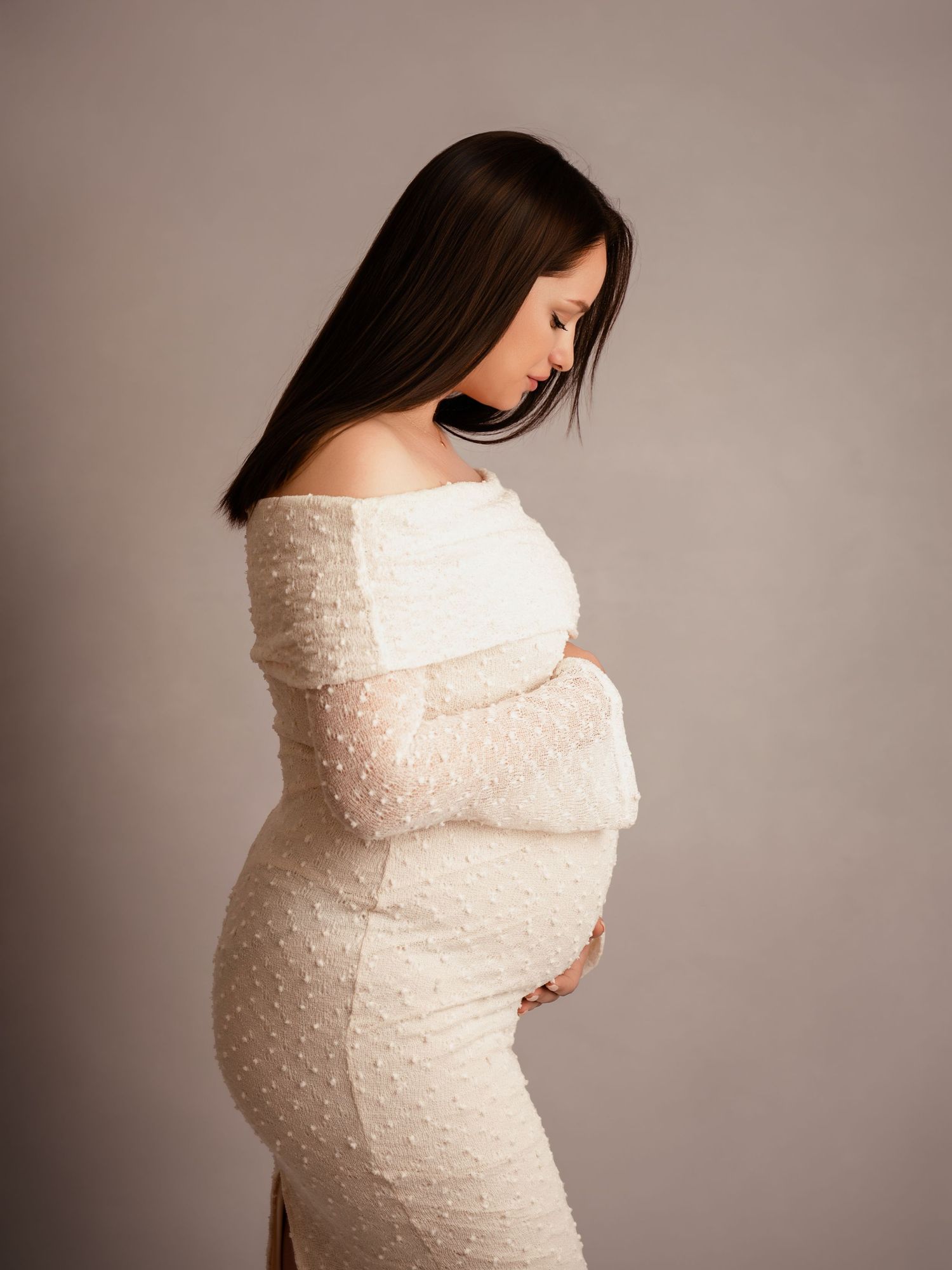 Pregnant woman in white lace off-shoulder gown cradling baby bump, side profile maternity photo studio portrait.
