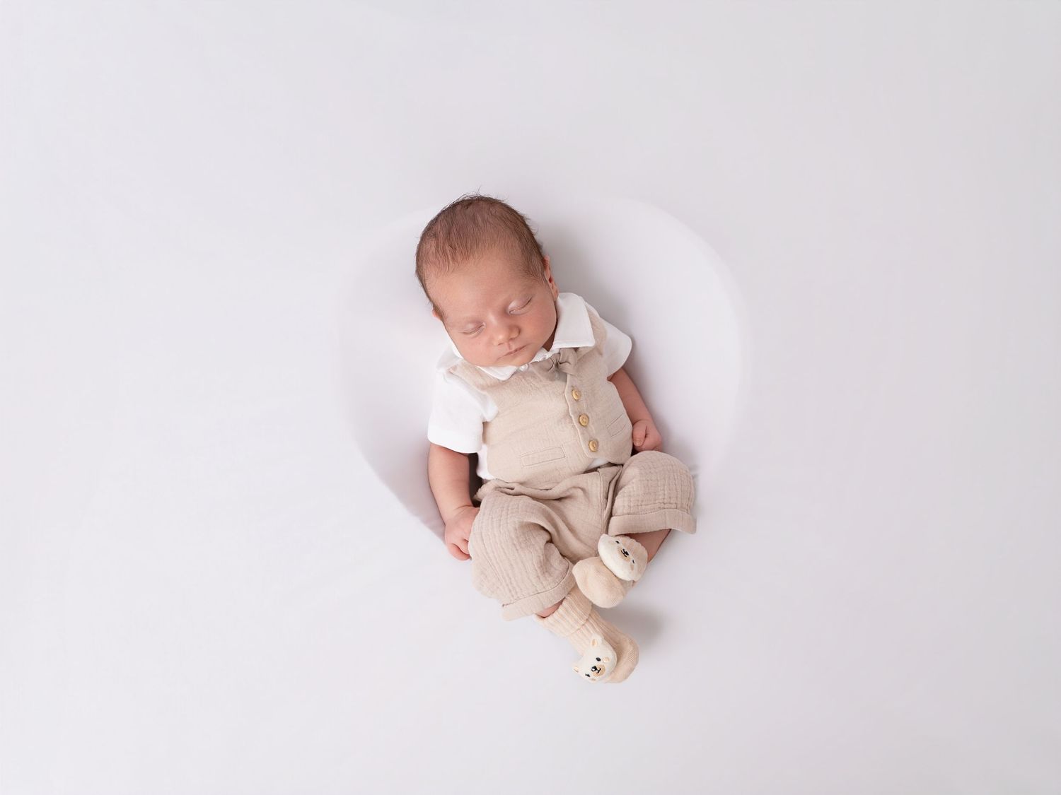 Newborn baby sleeping peacefully in beige overalls and white shirt on a soft white background.