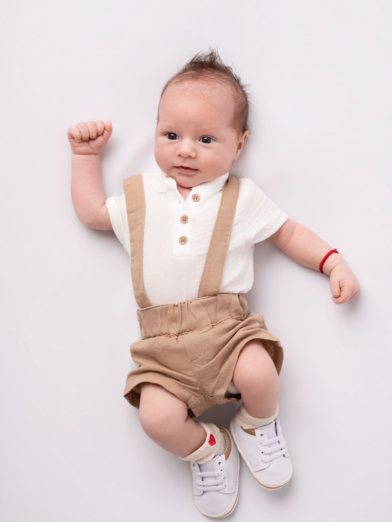 Adorable baby boy in white polo shirt and tan suspender shorts lying on white background with fist raised.