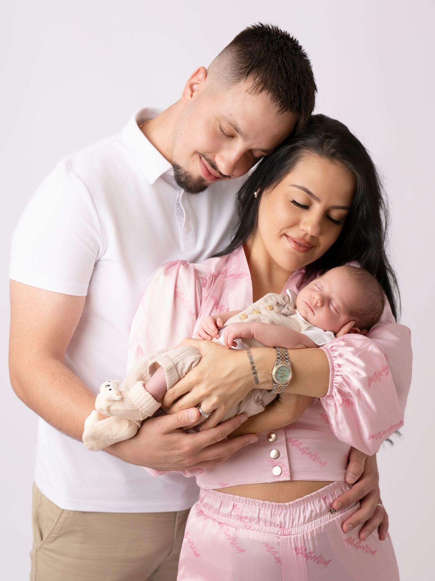 Happy young parents in pink outfits tenderly hold their sleeping newborn baby in a bright studio photo.