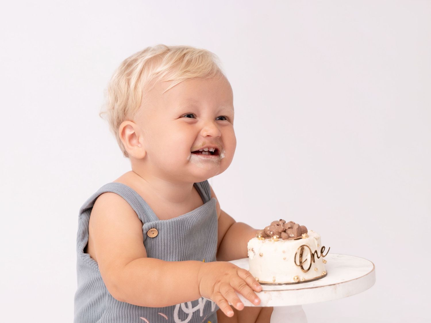 Smiling toddler boy in gray romper holding a small white first birthday smash cake with gold One topper.